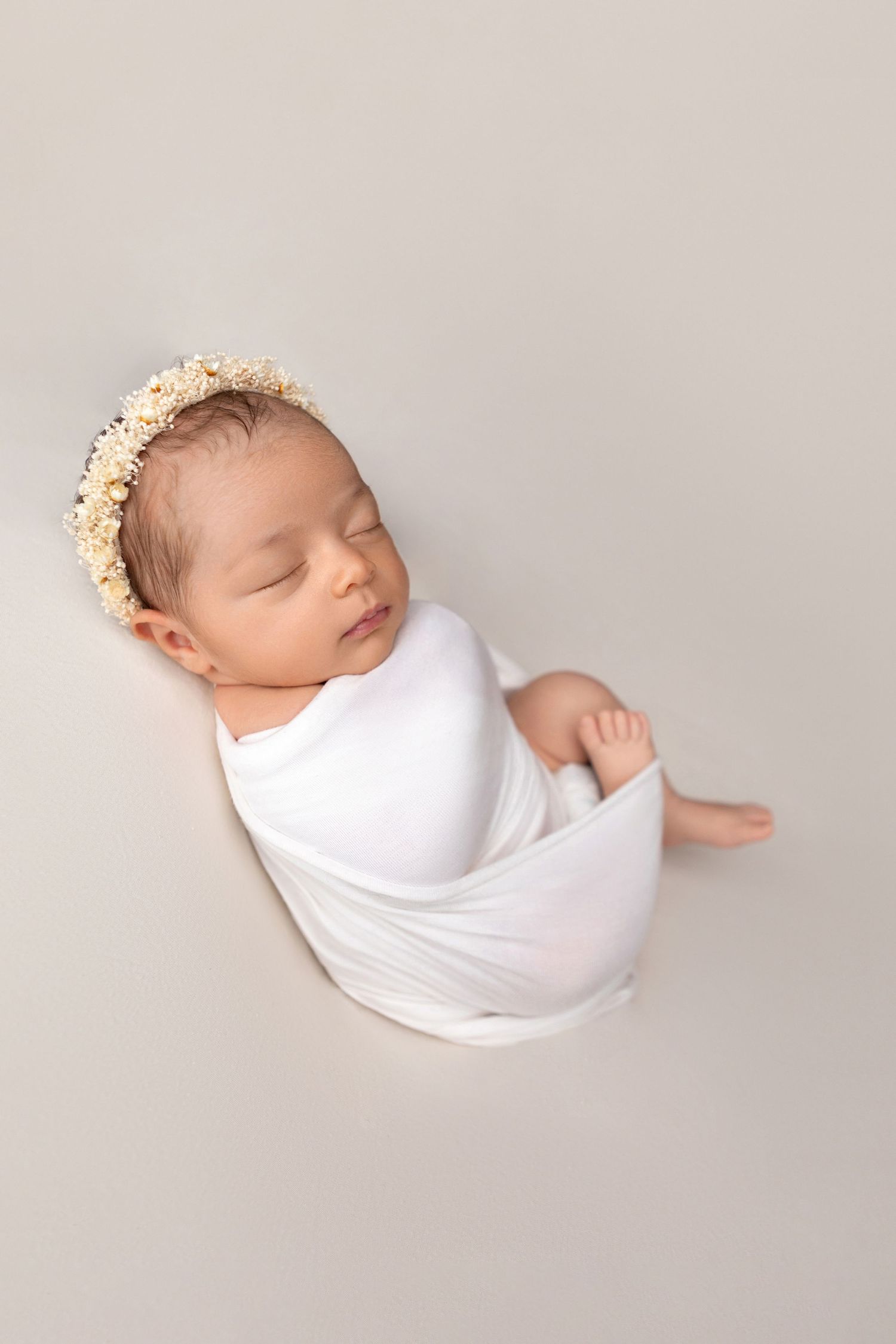 Peaceful newborn baby wrapped in white swaddle cloth with a cream floral headband, photographed from above.