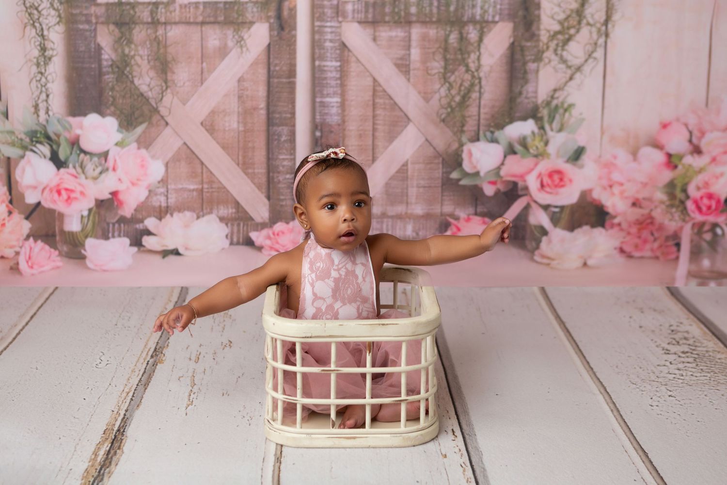 Adorable baby girl in pink lace romper sitting in white basket with floral barn door backdrop.