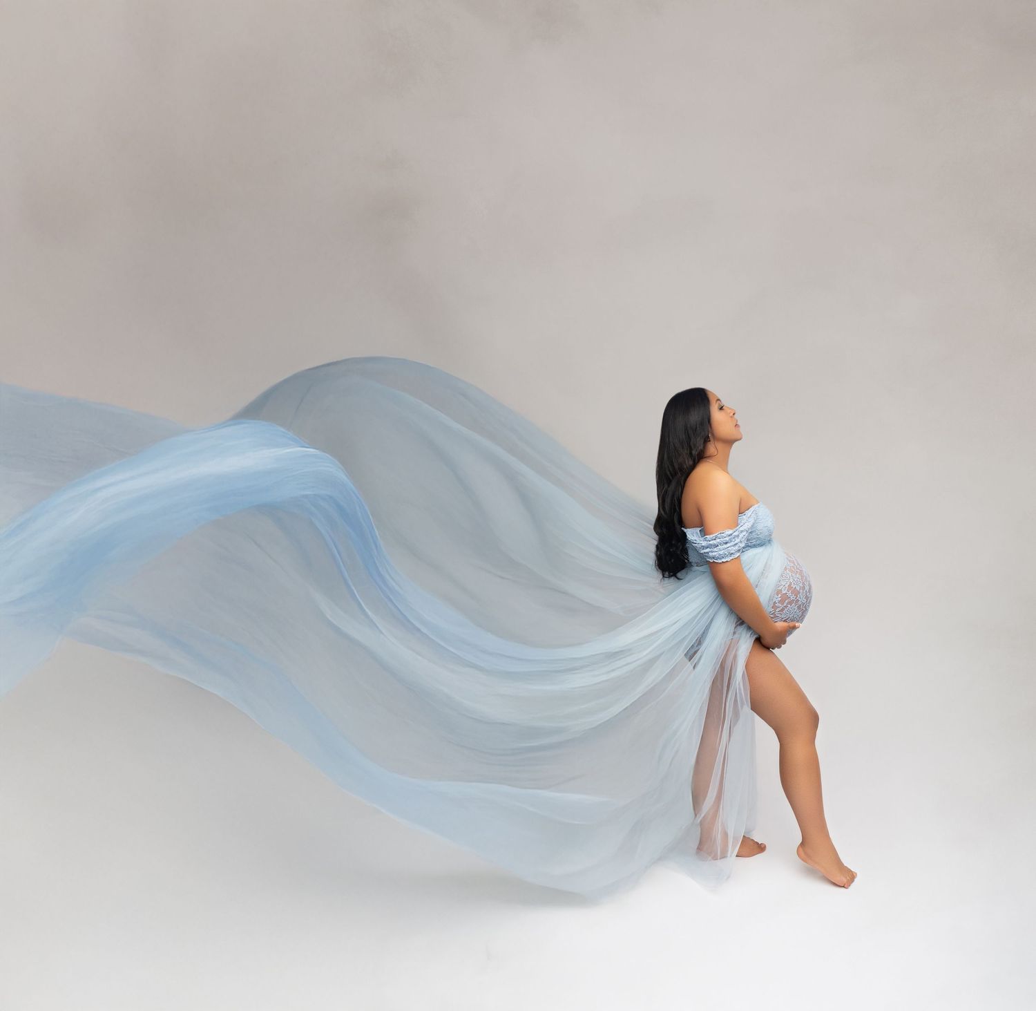 Pregnant woman in flowing light blue chiffon gown posing gracefully in a studio maternity photoshoot.
