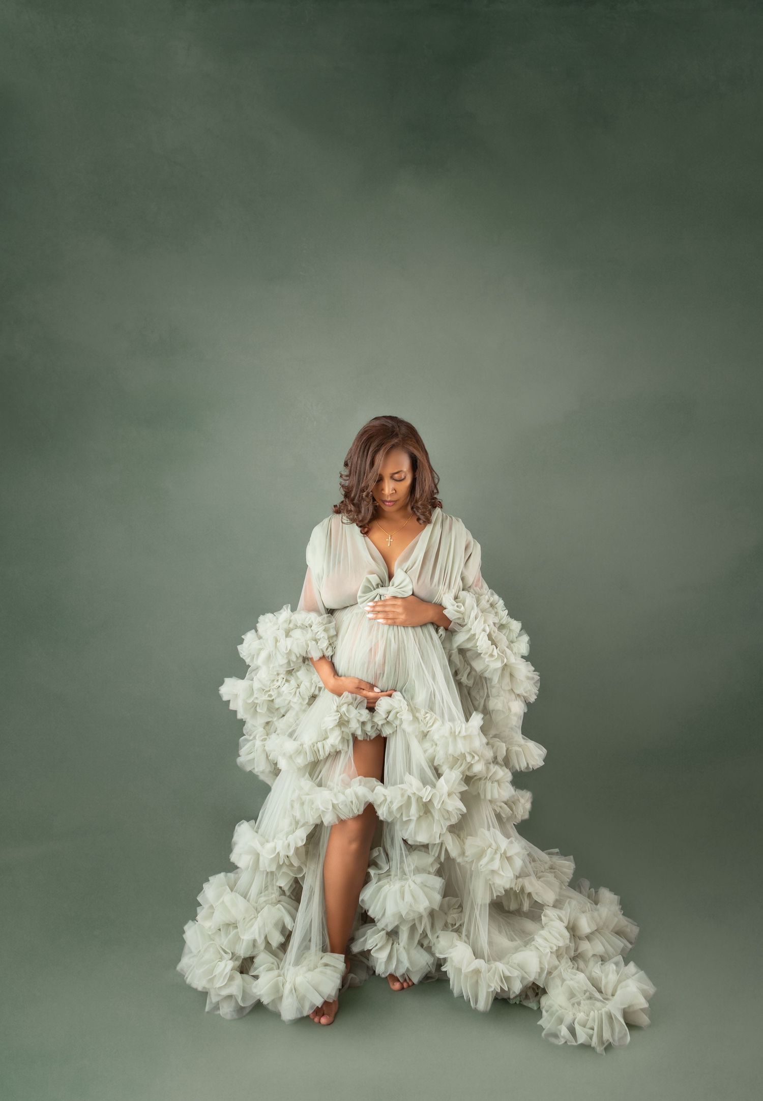 Pregnant woman in white ruffled maternity gown cradling bump, studio portrait with sage green background.