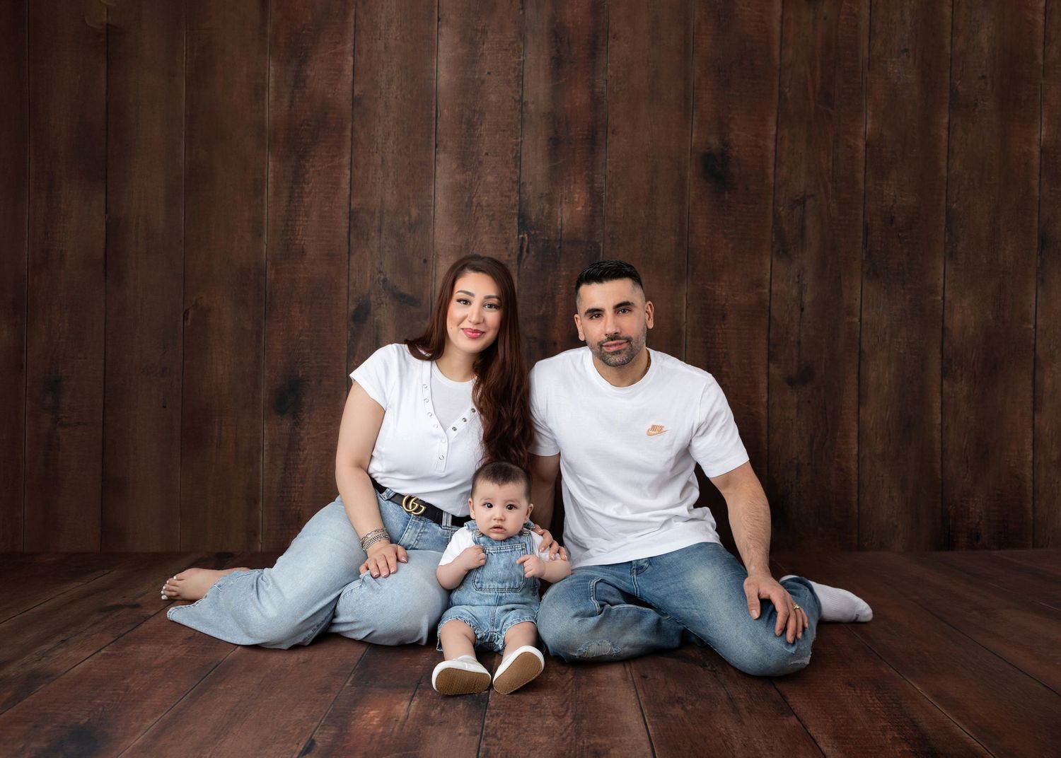 Young family with baby sitting on wood floor against rustic dark wood wall backdrop in matching white tops.