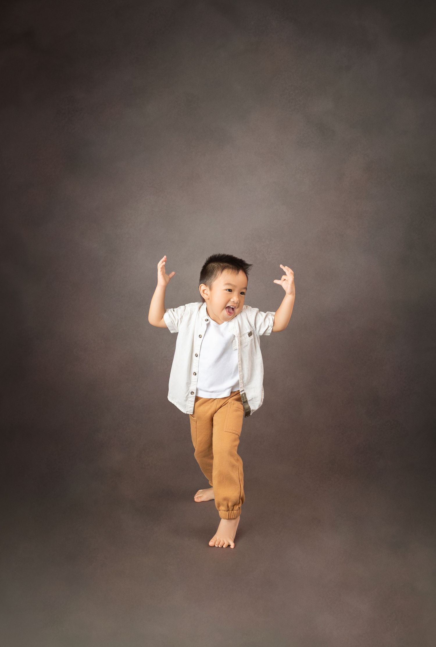 Energetic young boy in white shirt and tan pants dancing barefoot against a gray studio background.