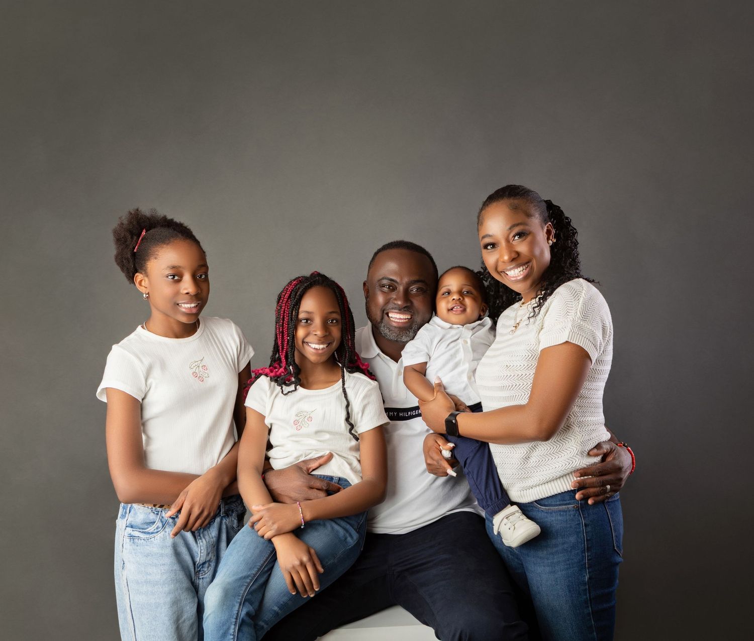 Happy family of five posing together in white shirts and jeans against a gray studio background.