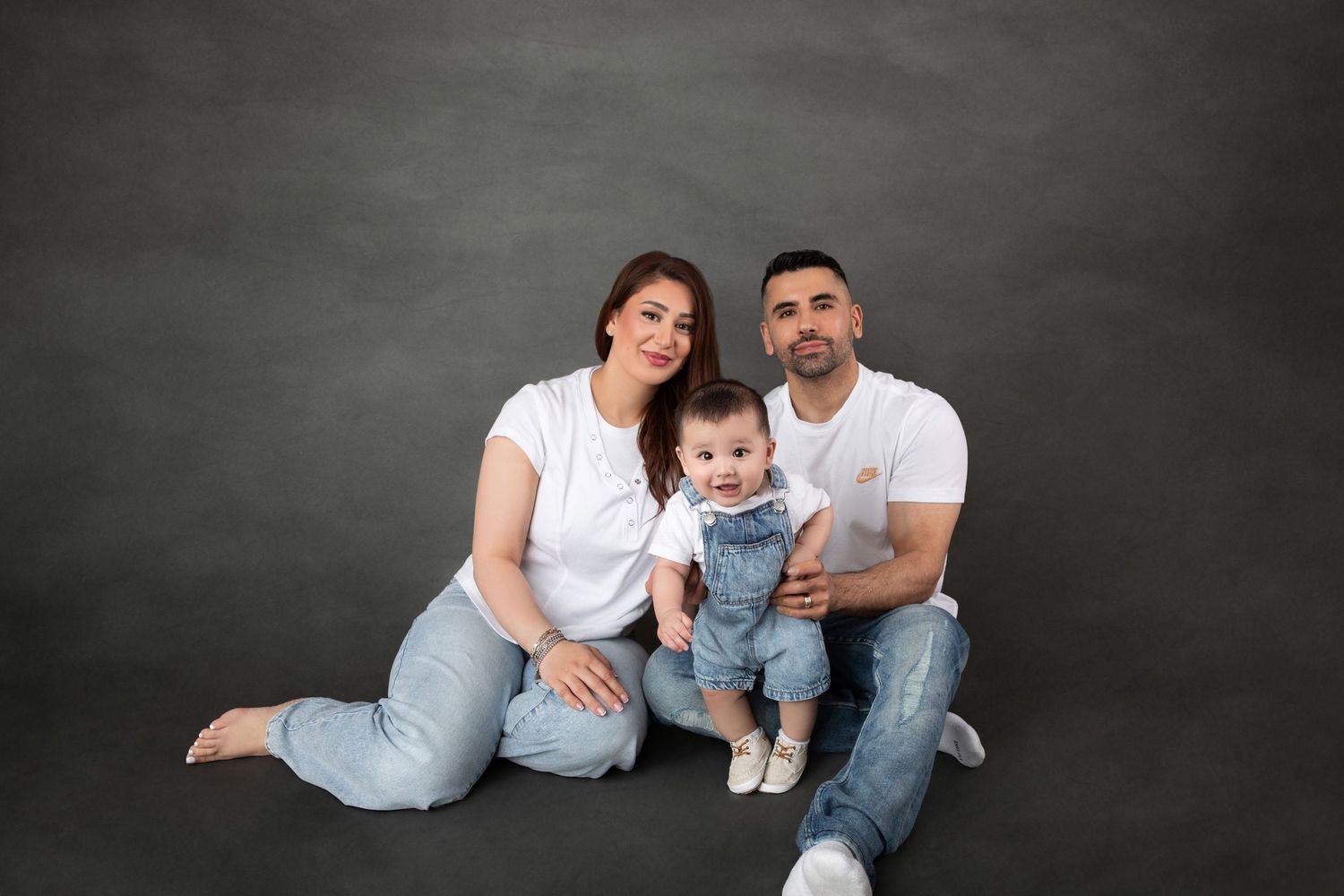 Family of three sitting together with baby in denim overalls on grey backdrop.