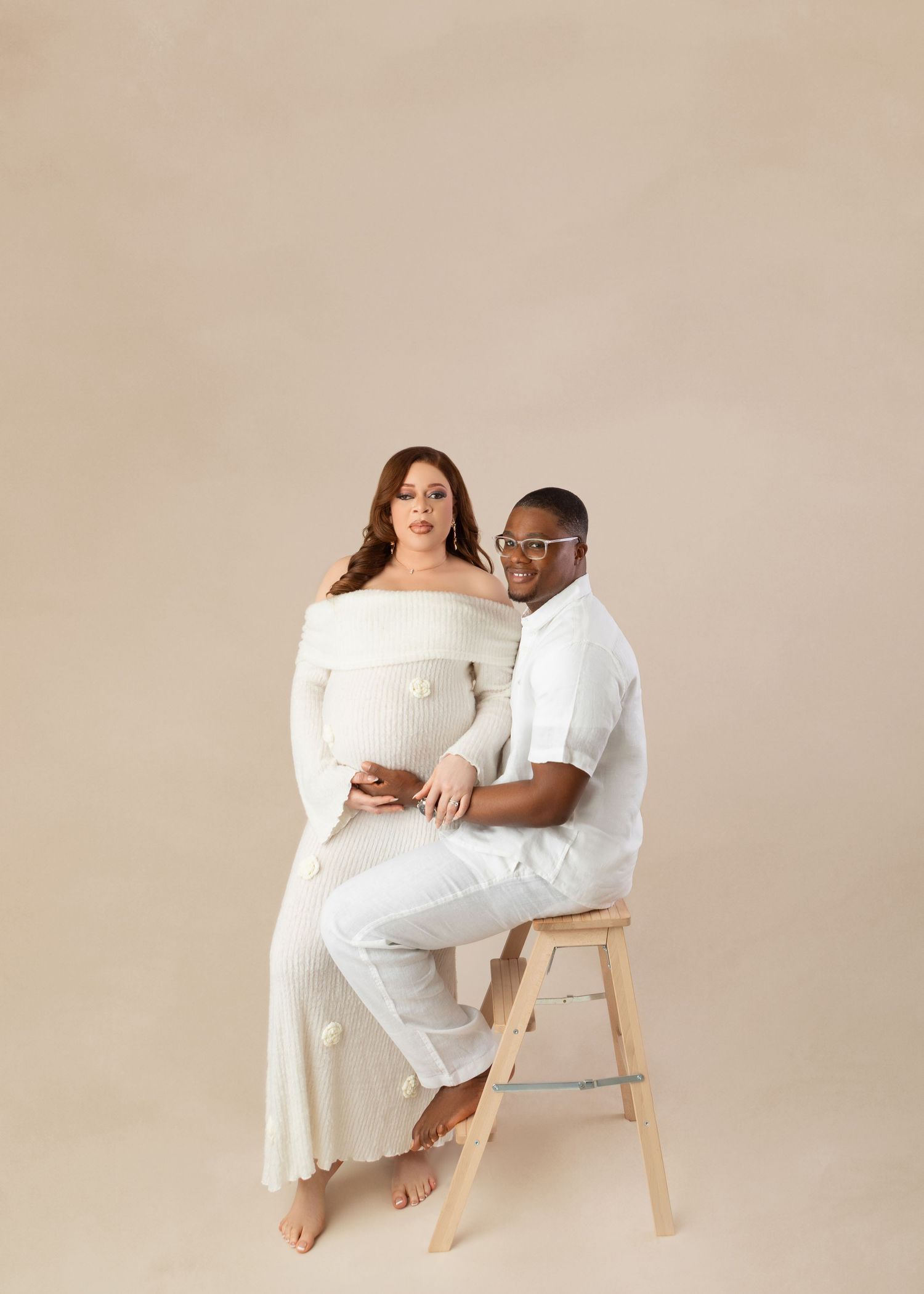 Couple posing for maternity photo on stool against beige backdrop, woman in white off-shoulder gown, man in white shirt.