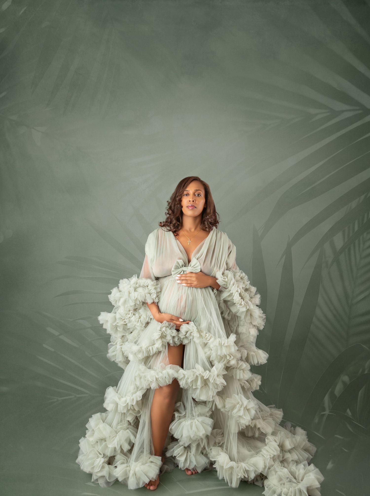 Pregnant woman in white ruffled maternity gown poses gracefully against a green tropical leaf backdrop.
