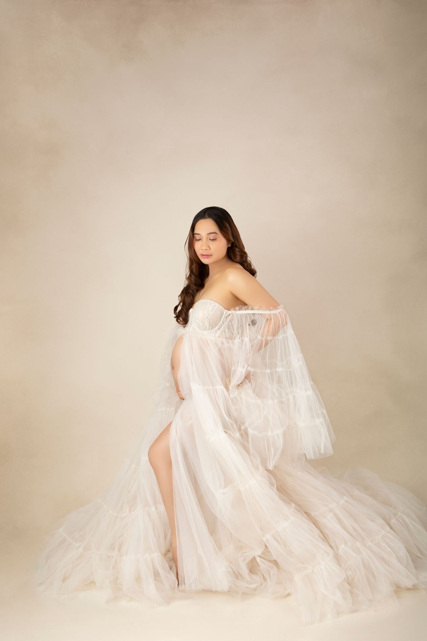 A pregnant woman in an elegant white off-shoulder tulle maternity gown poses on a beige background.