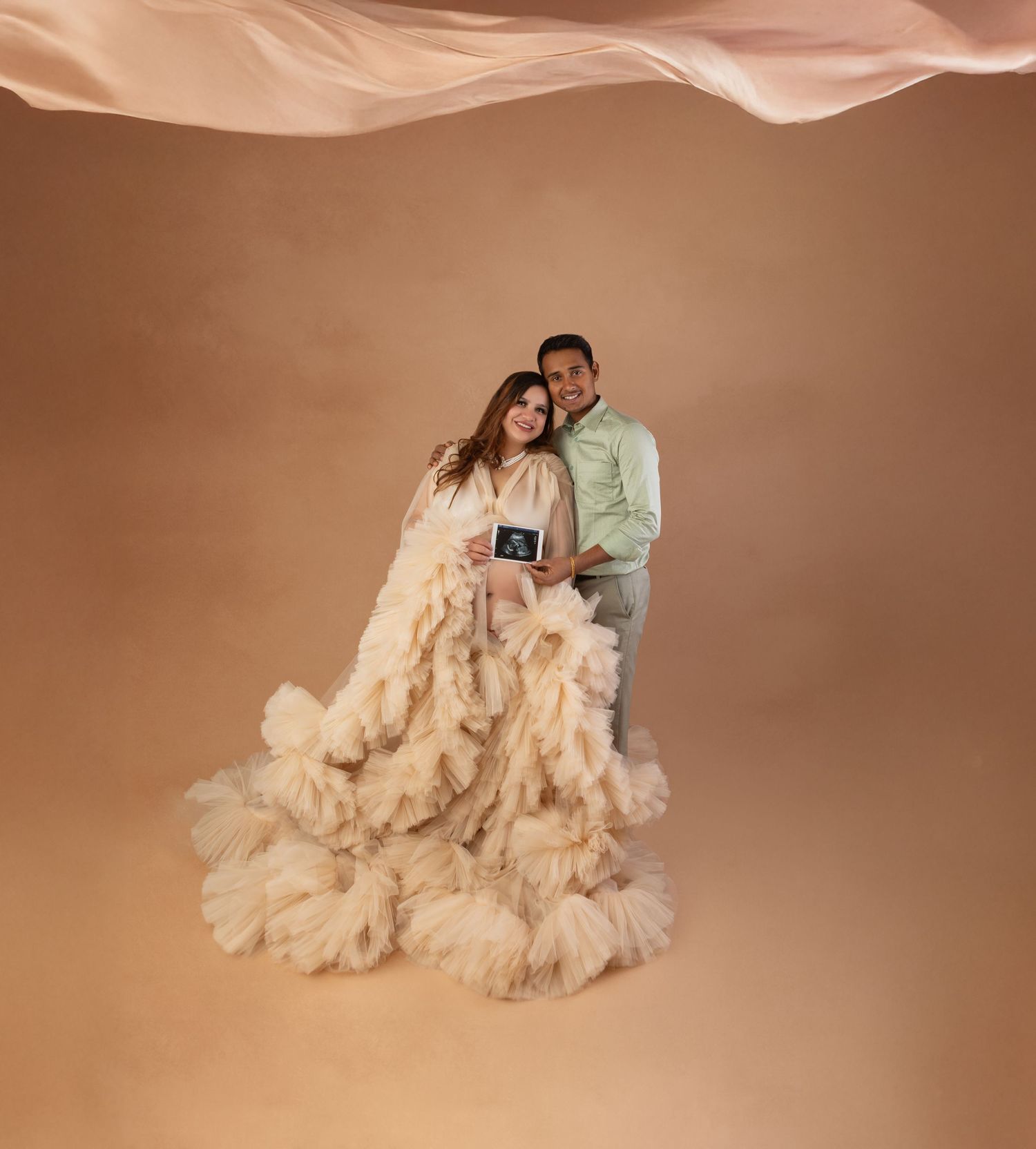 Expecting couple in cream tulle gown holding ultrasound photo in warm-toned professional maternity studio photo.