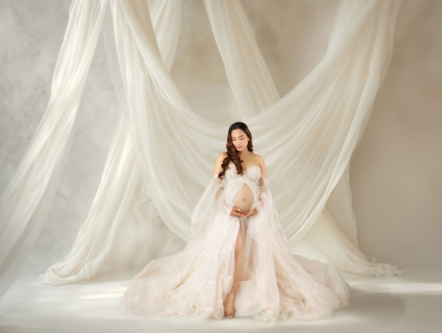 Pregnant woman in elegant off-shoulder blush gown poses gracefully against flowing white drapes in a studio.