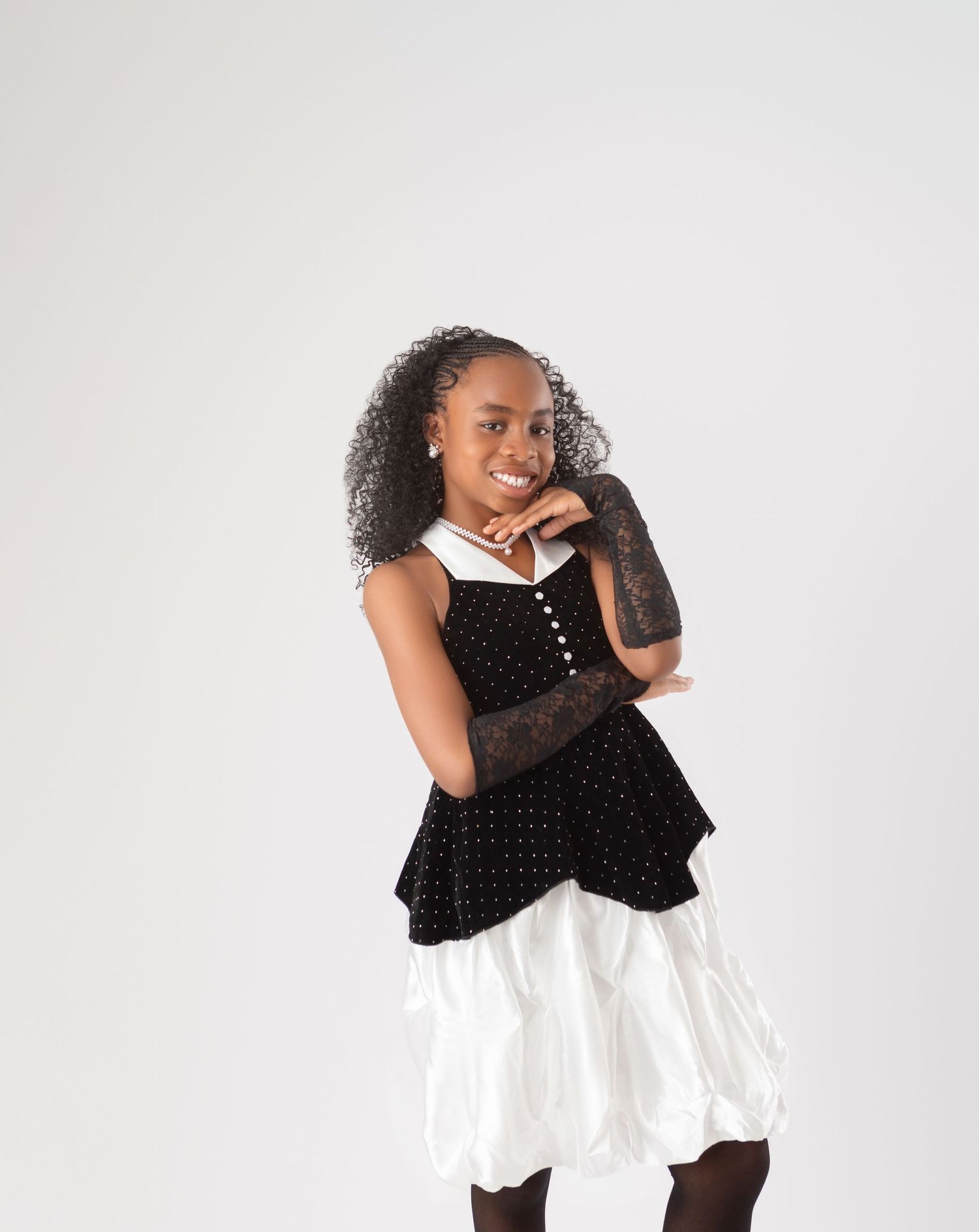 Young girl smiling in black velvet top, white bubble skirt, and lace gloves against a light gray background.