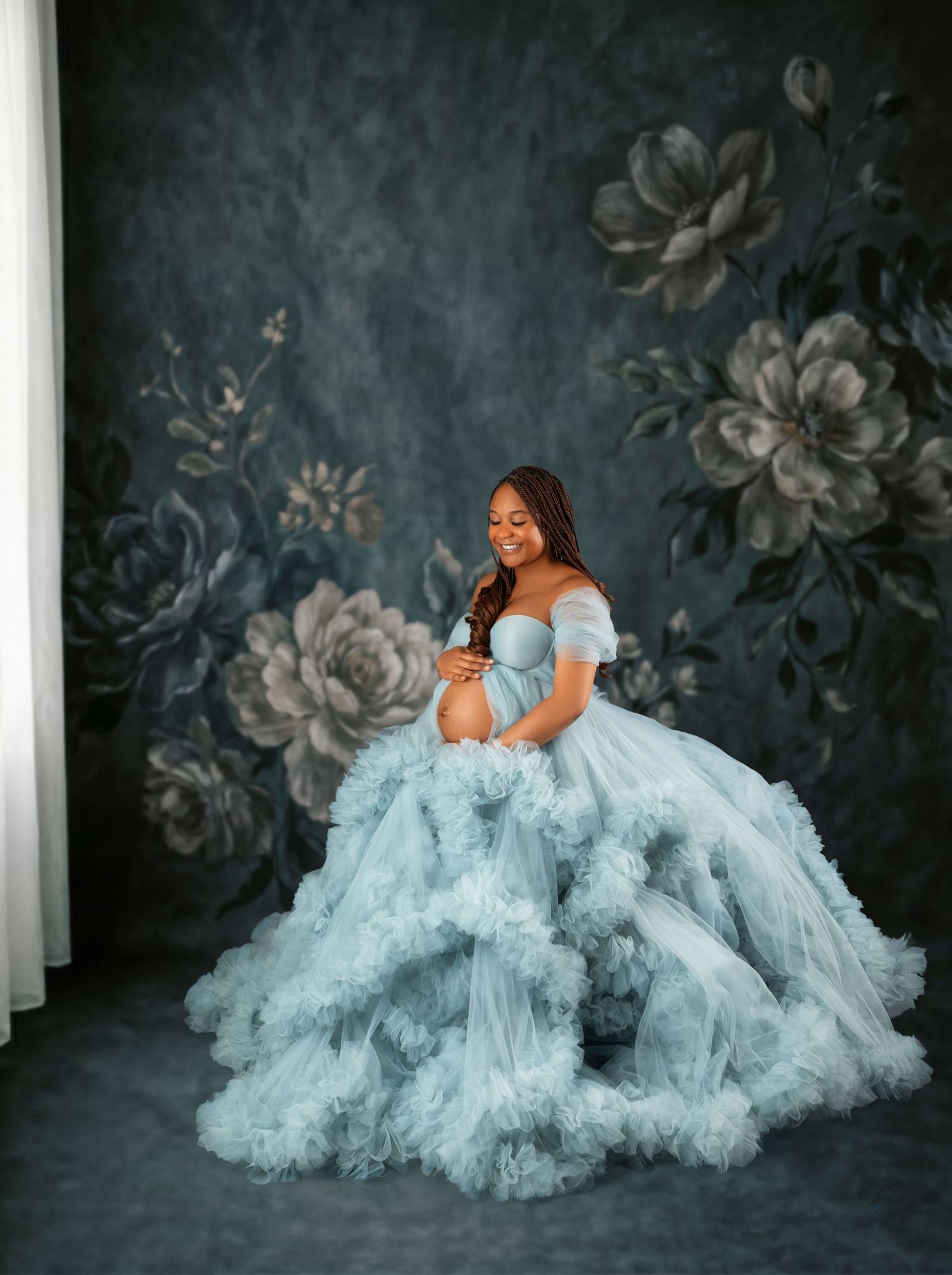 Pregnant woman in a flowing light blue tulle gown poses gracefully against a dark floral backdrop in a maternity photoshoot.