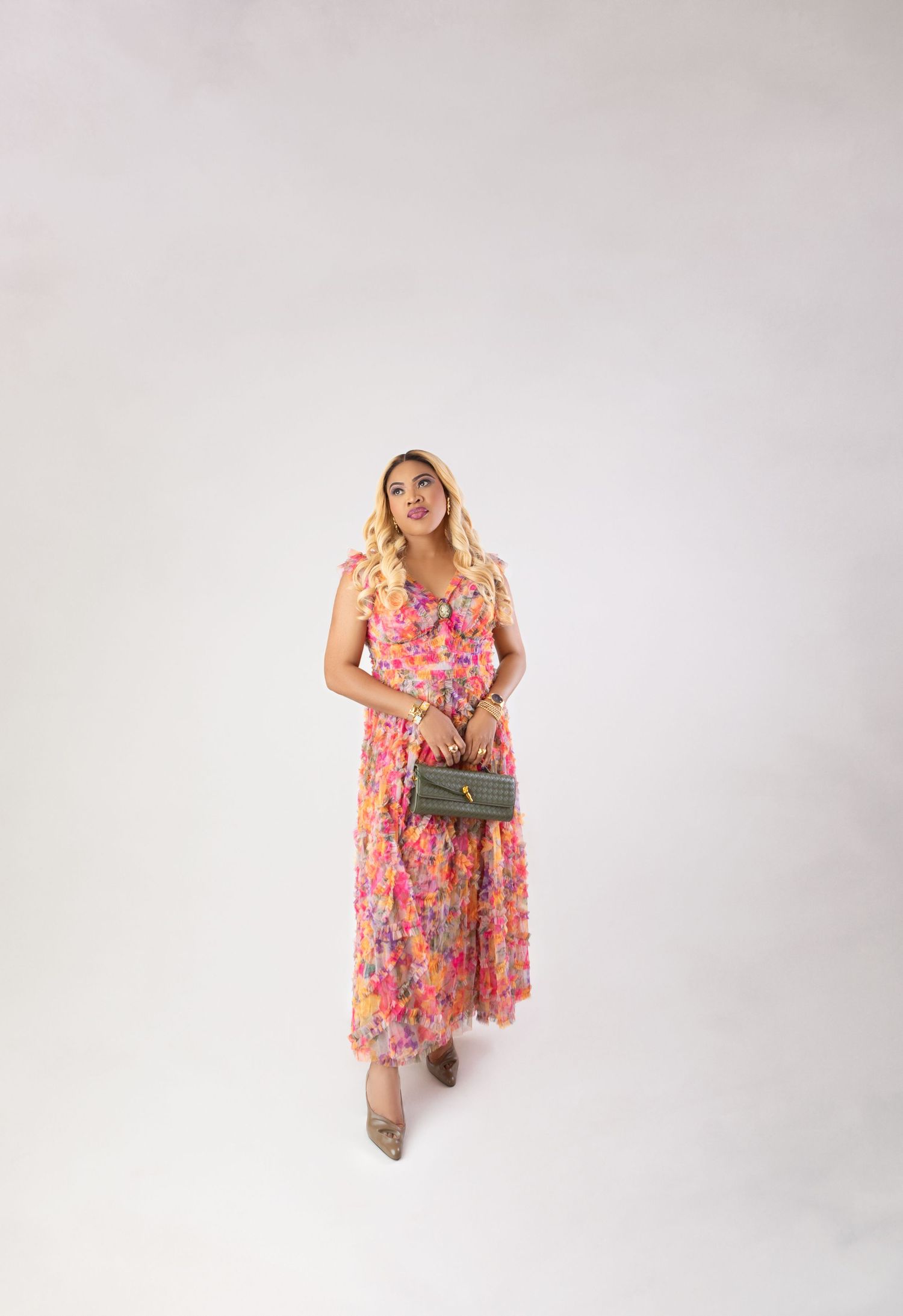 Woman in pink floral maxi dress holding olive green clutch bag, wearing nude heels, standing against white background.