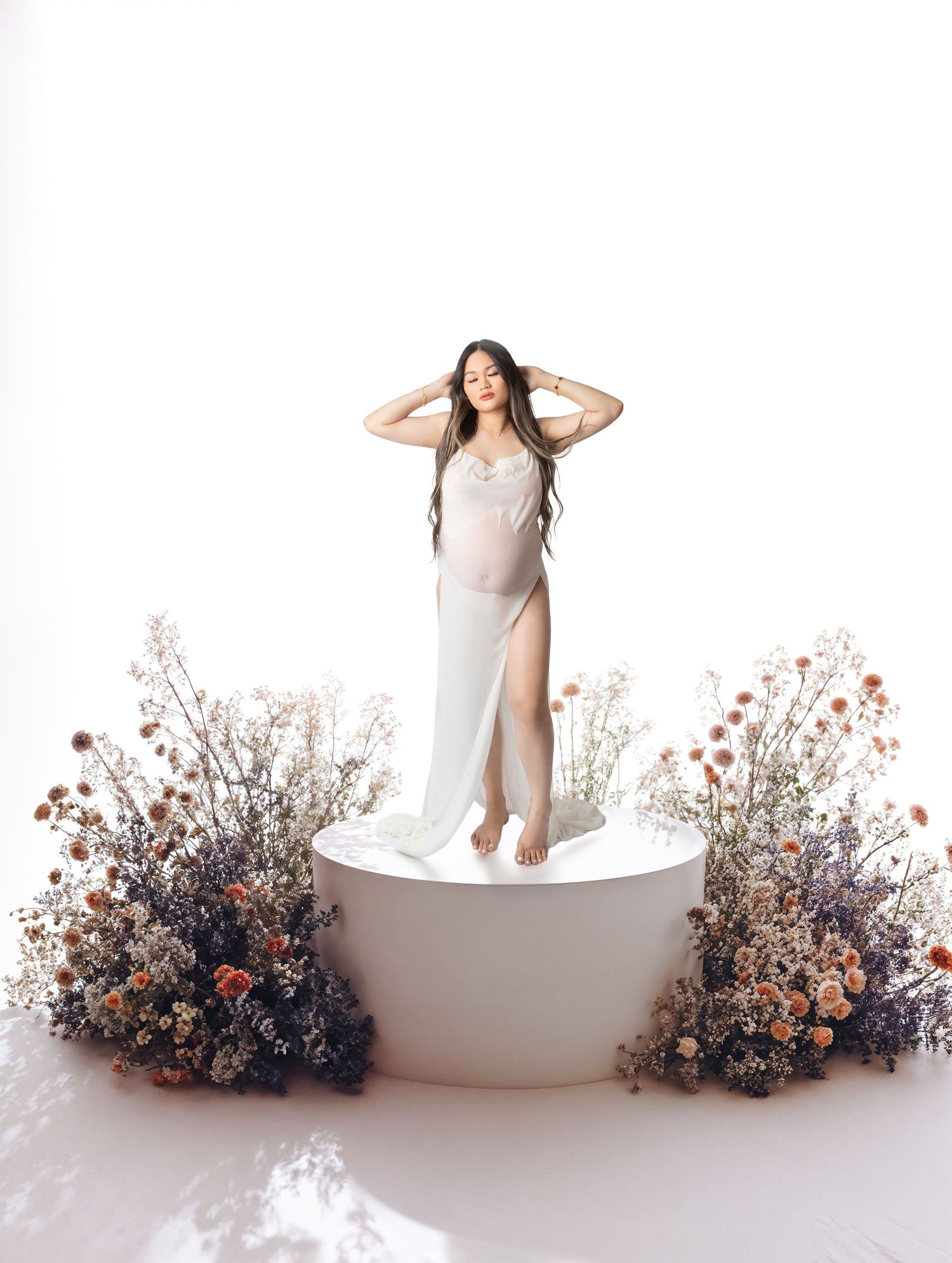 Pregnant woman in white gown standing on a round platform surrounded by wildflowers in bright studio.