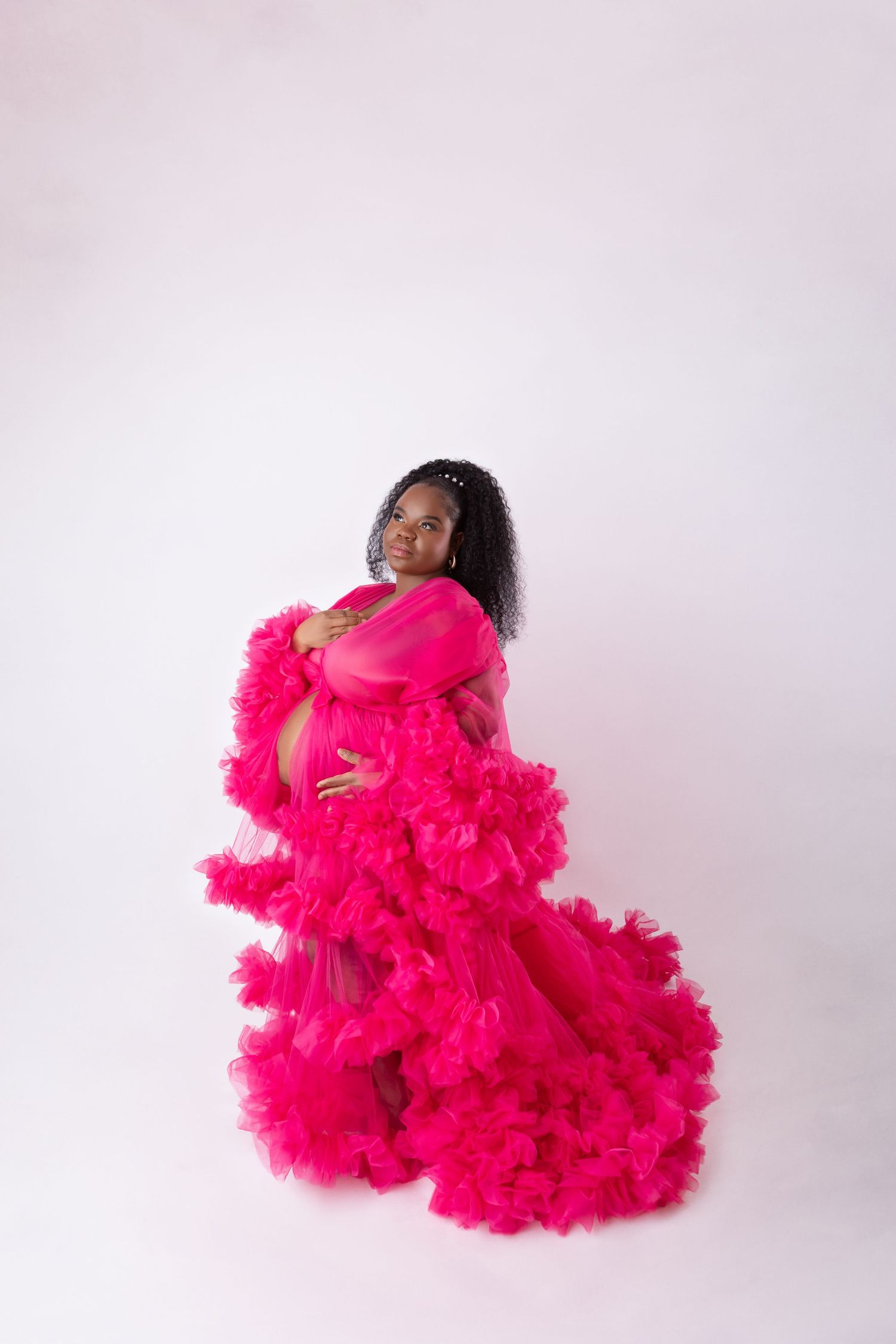 Pregnant woman in a stunning hot pink ruffled tulle maternity gown posing against a white background.