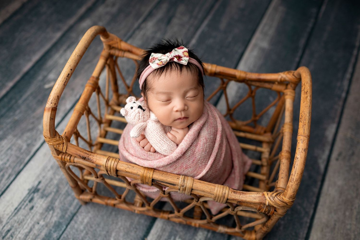Peacefully sleeping newborn wrapped in pink swaddle inside wicker basket with wooden floor backdrop.