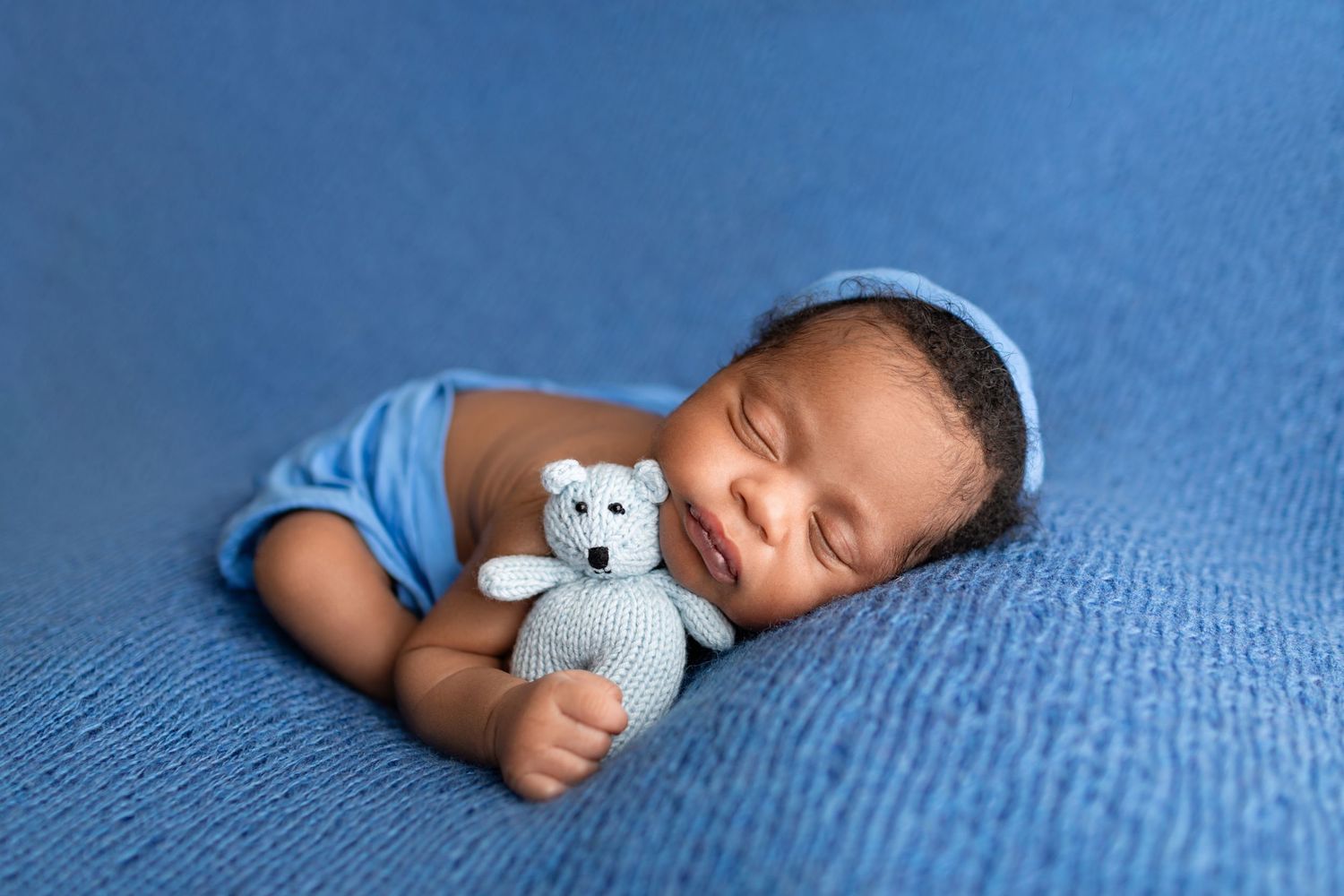 Sleeping newborn baby boy in blue knit hat holding a small teddy bear on a blue blanket.