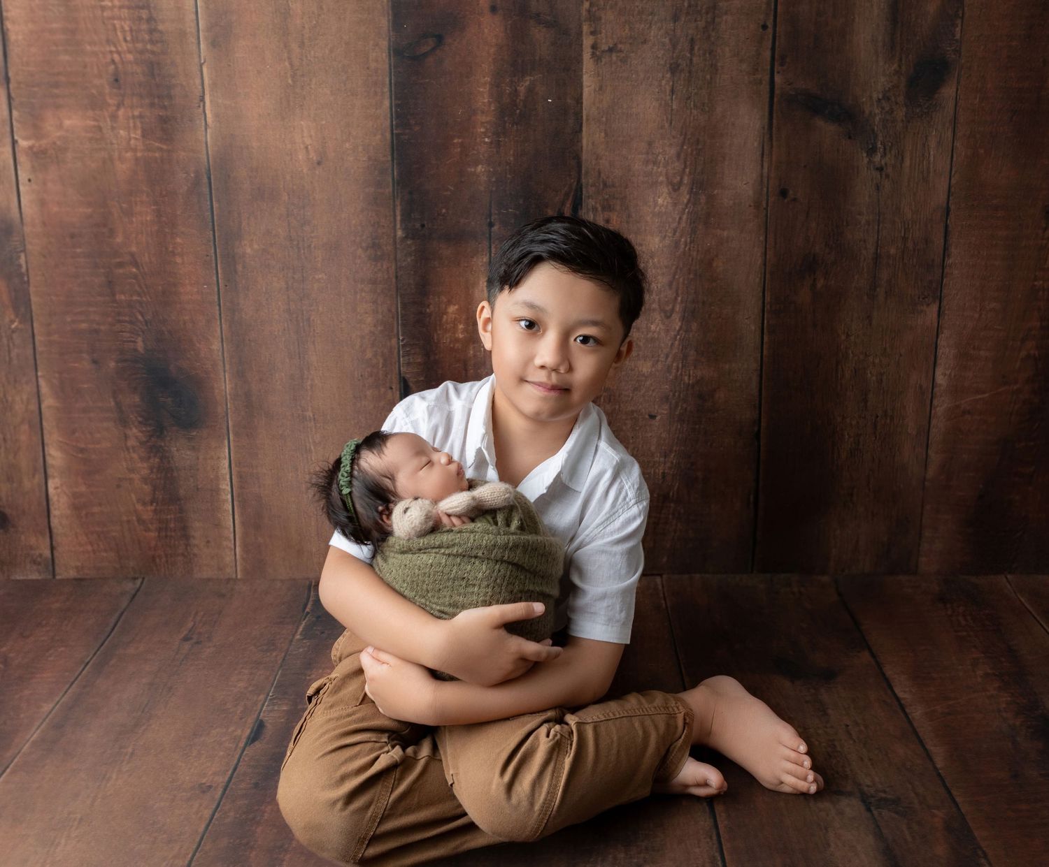 A young boy sits cross-legged holding a sleeping newborn wrapped in green against a rustic wood backdrop.