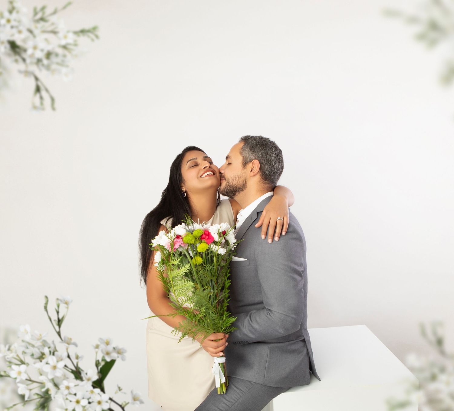 Smiling couple sharing a romantic kiss, woman holding colorful flower bouquet in white studio setting.