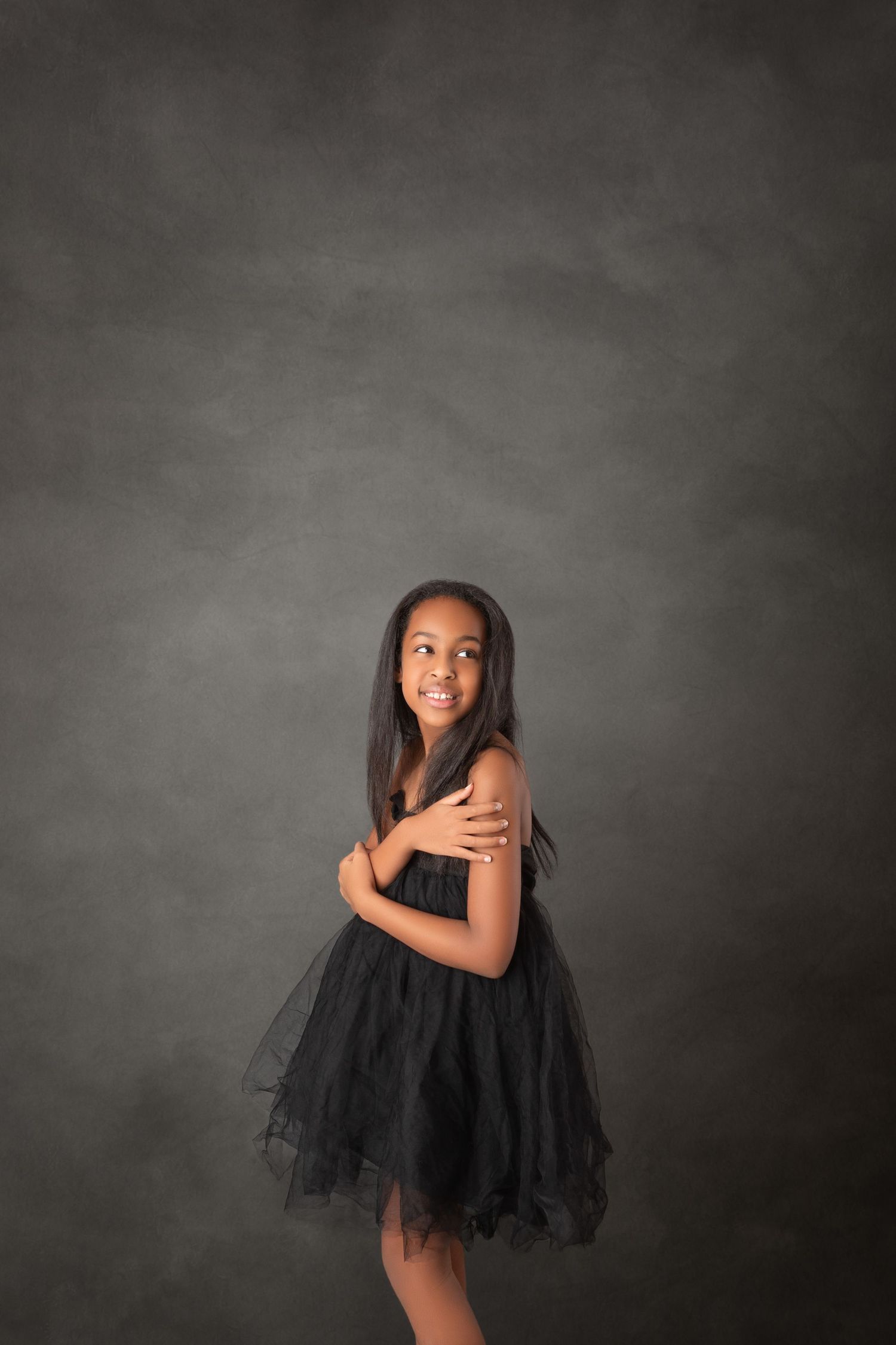 Young girl in black tutu dress poses gracefully against gray backdrop, smiling over shoulder with arms crossed.