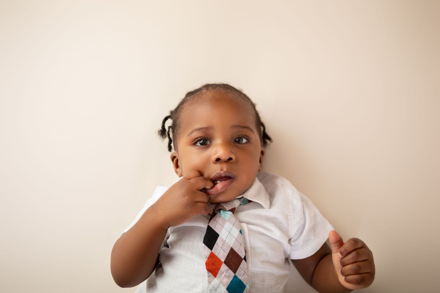Adorable toddler in white shirt and colorful argyle tie posing with finger in mouth against neutral background.