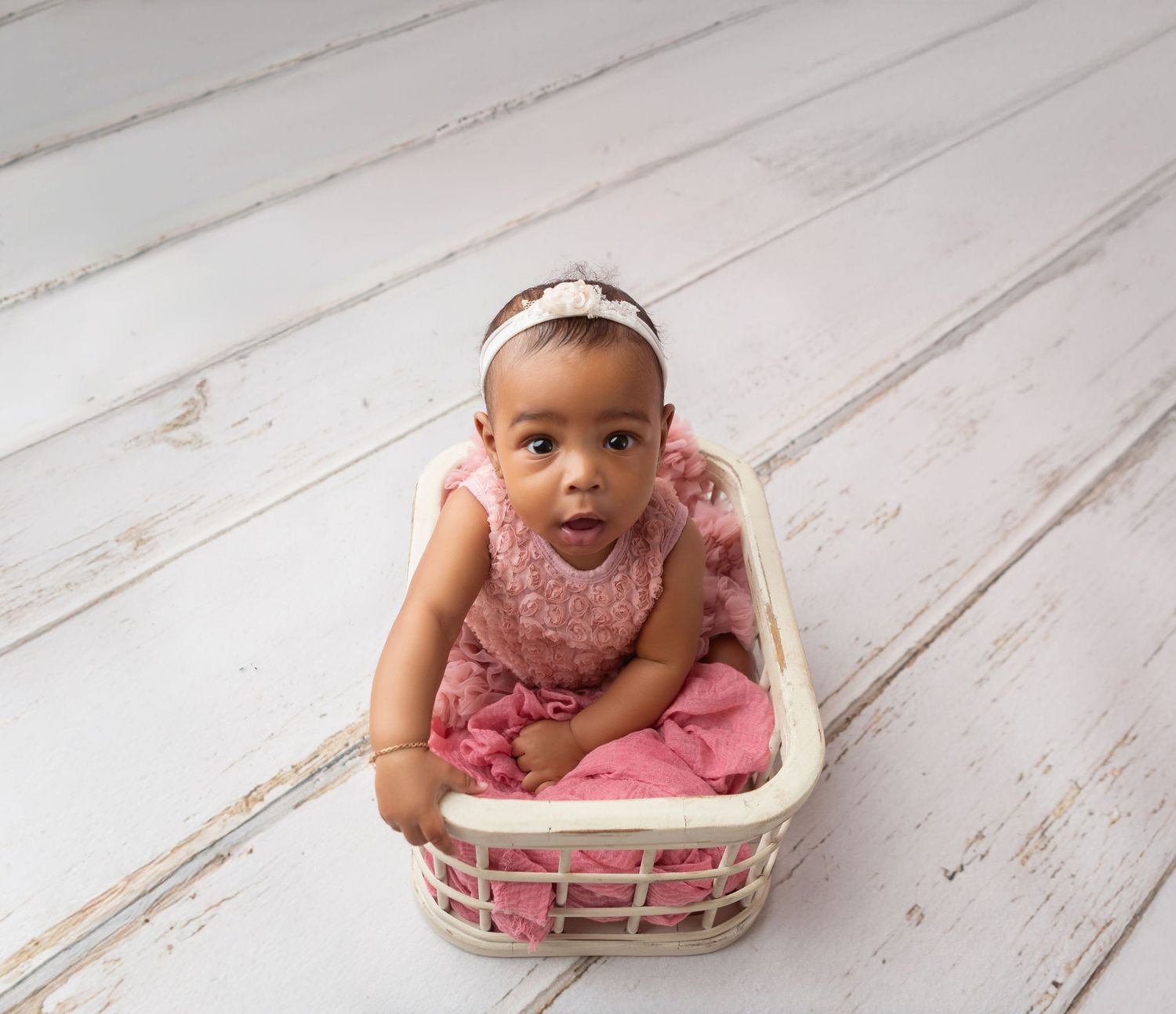 Precious baby girl in pink lace romper sitting in white wicker basket on whitewashed floor.