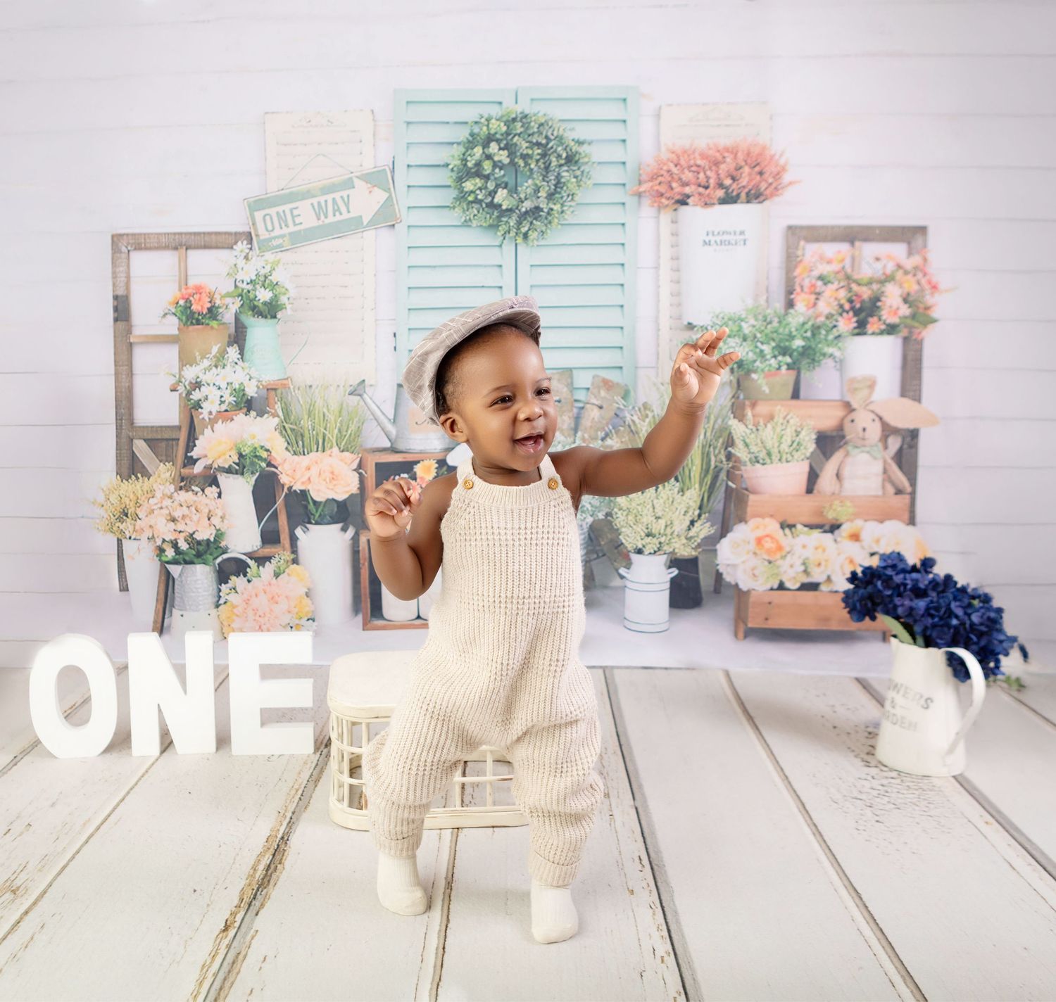 Adorable baby in cream overalls celebrates first birthday with floral backdrop and ONE sign.