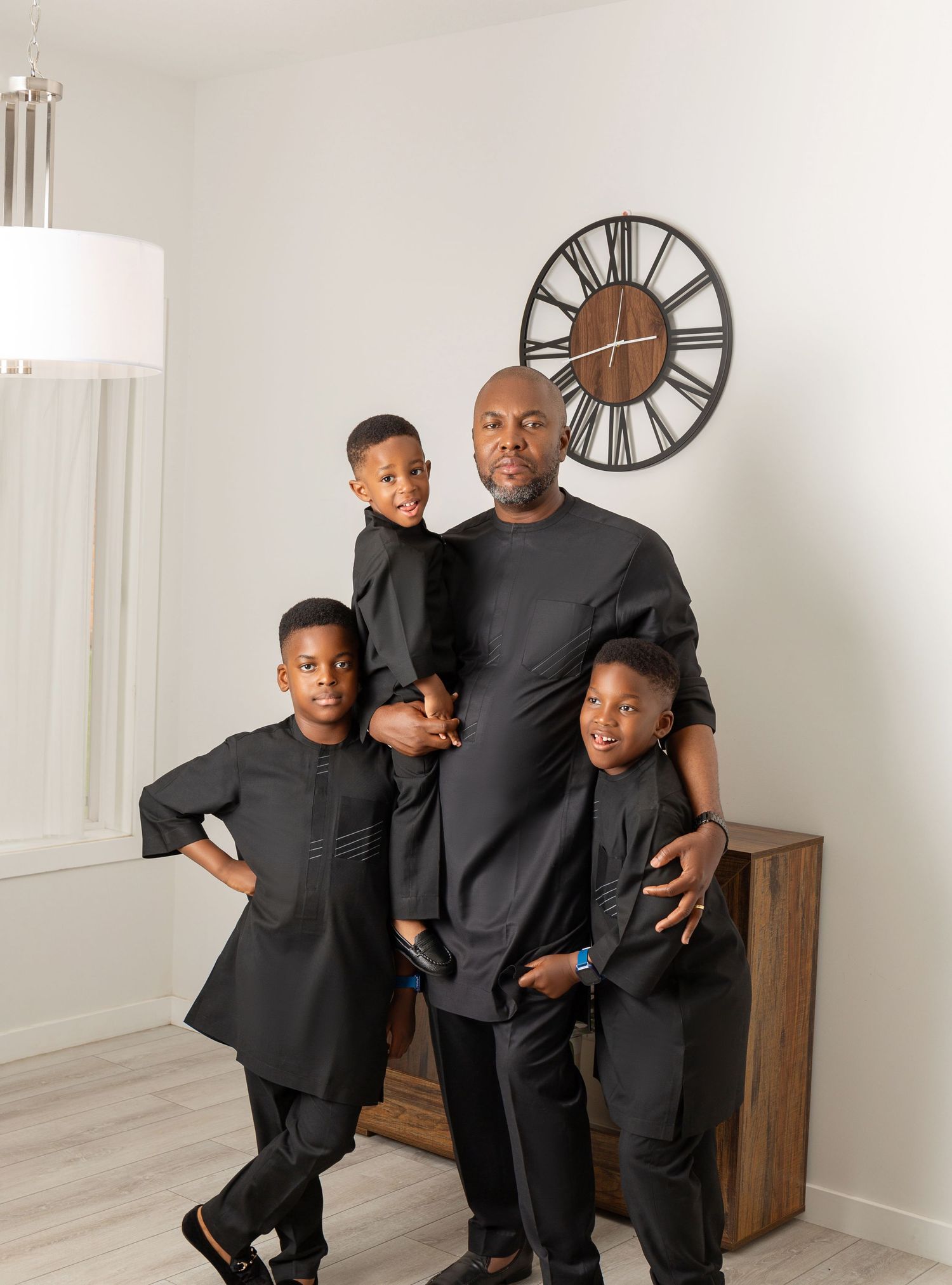 A father and three children pose in matching black outfits in a bright modern room with a decorative wall clock.