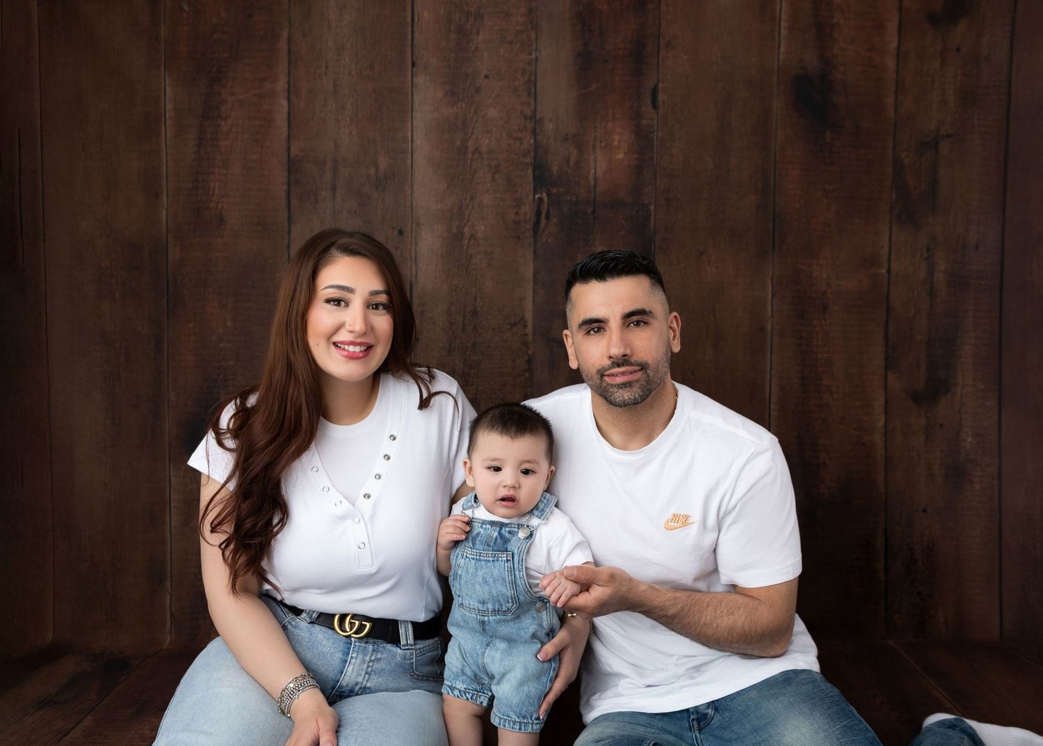 A young family with a baby boy poses for a portrait against a rustic wood backdrop, wearing matching white tops and jeans.