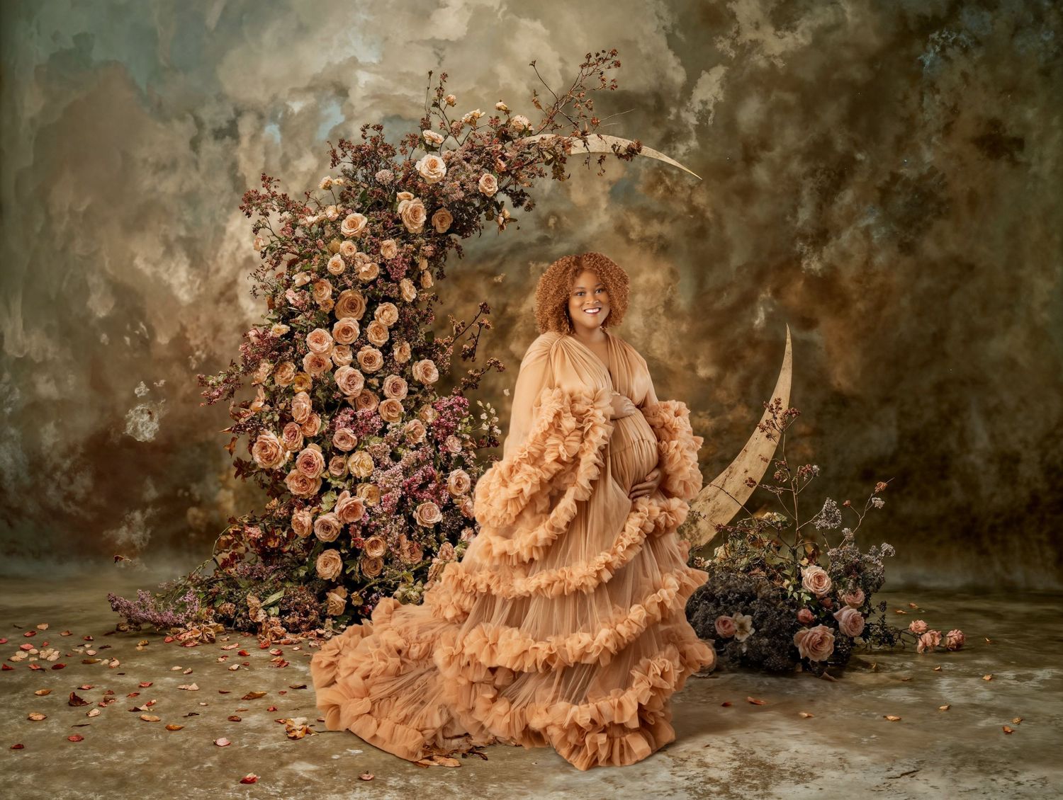 Woman in peach ruffled gown poses with floral crescent moon arrangement against painted backdrop.