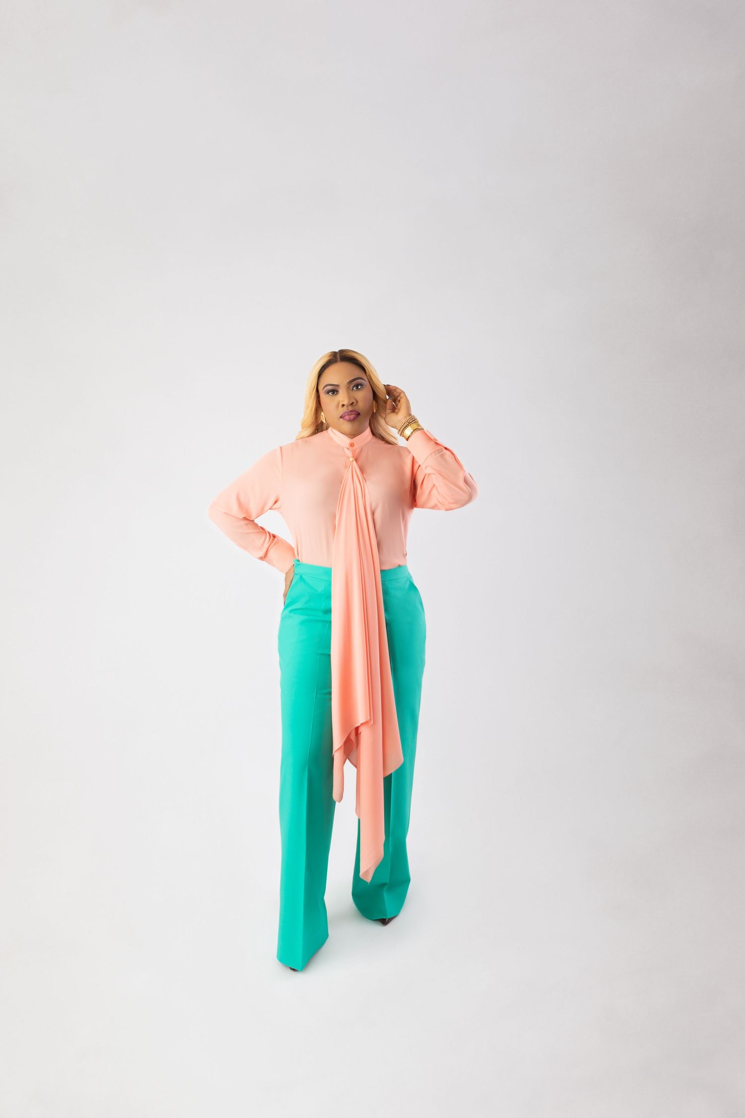 Woman in pink blouse with draped tie and teal wide-leg pants posing against white background.