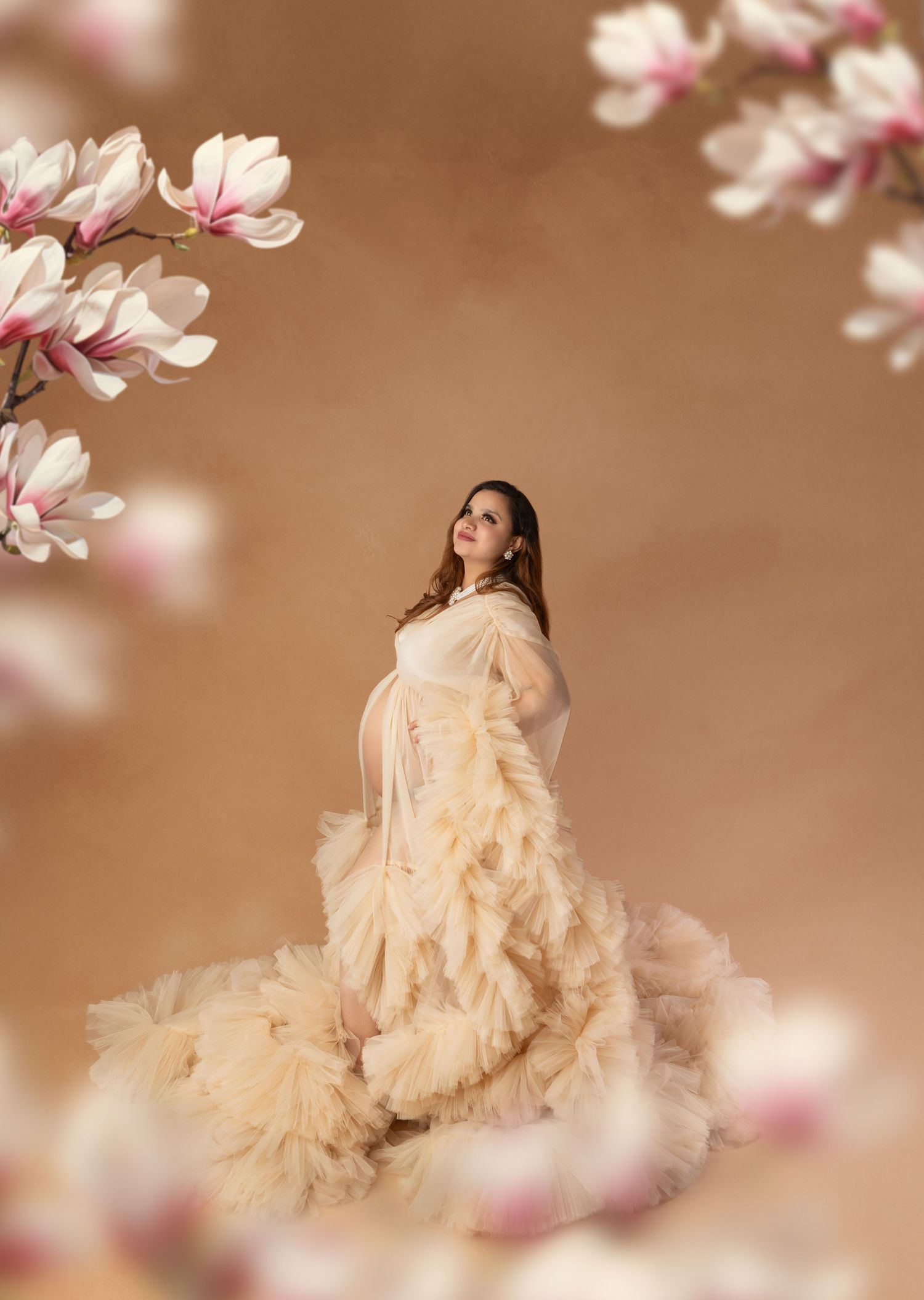 Pregnant woman in cream ruffle tulle gown surrounded by pink magnolia blossoms in elegant maternity studio portrait.