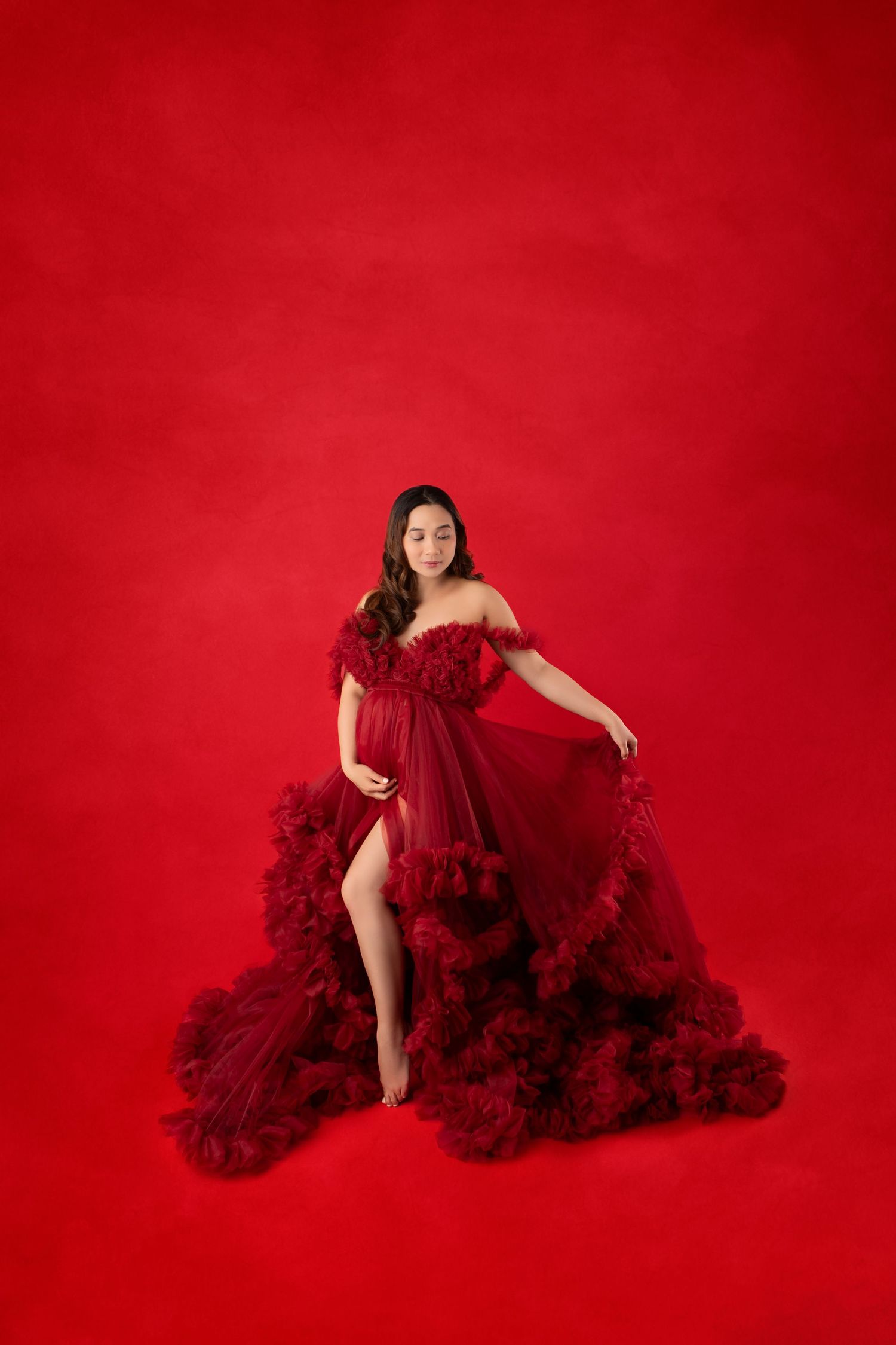 Pregnant woman in elegant dark red ruffled tulle gown posing gracefully against a rich red studio background.