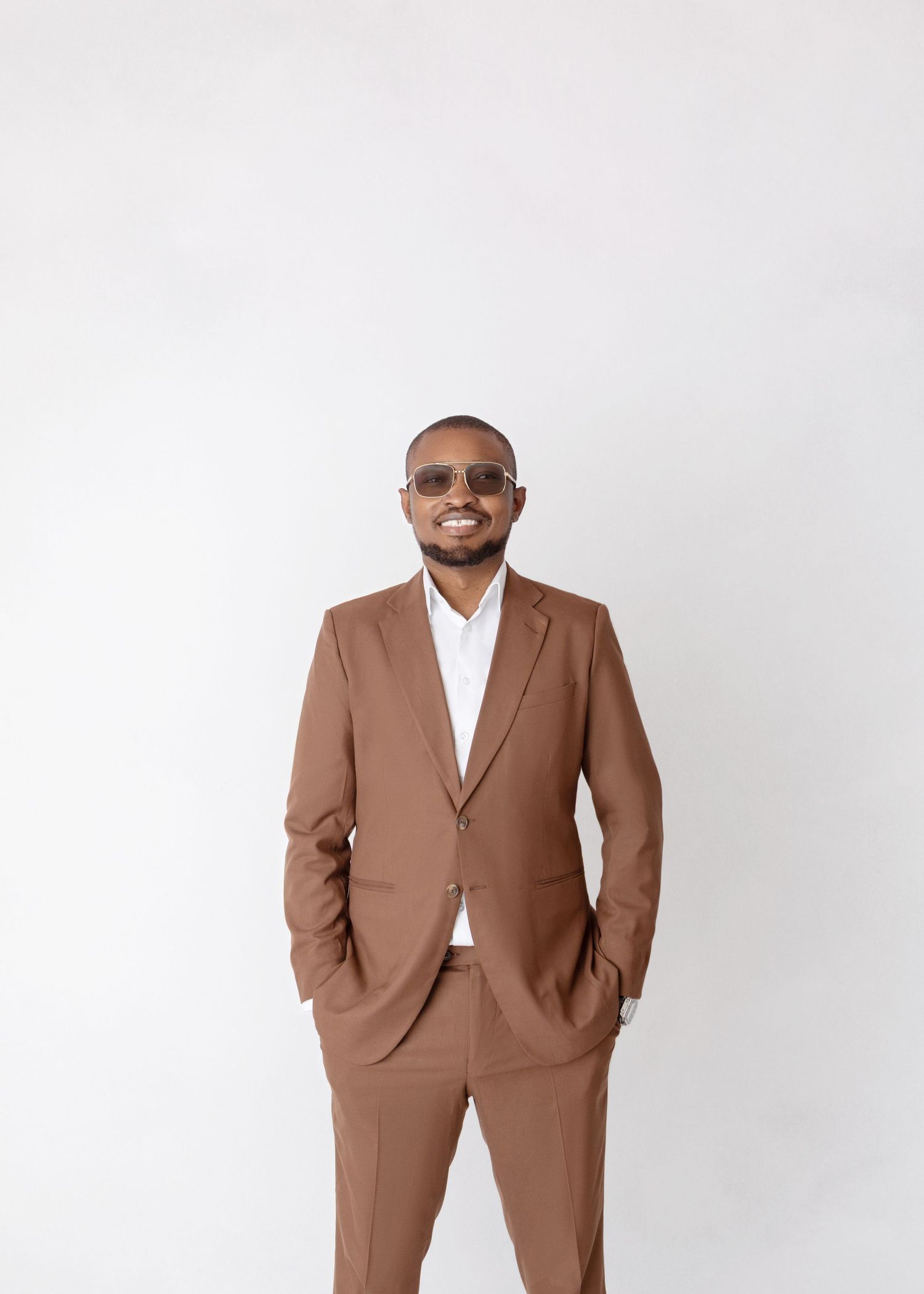 Smiling man in brown suit and sunglasses posing with hands in pockets in studio.