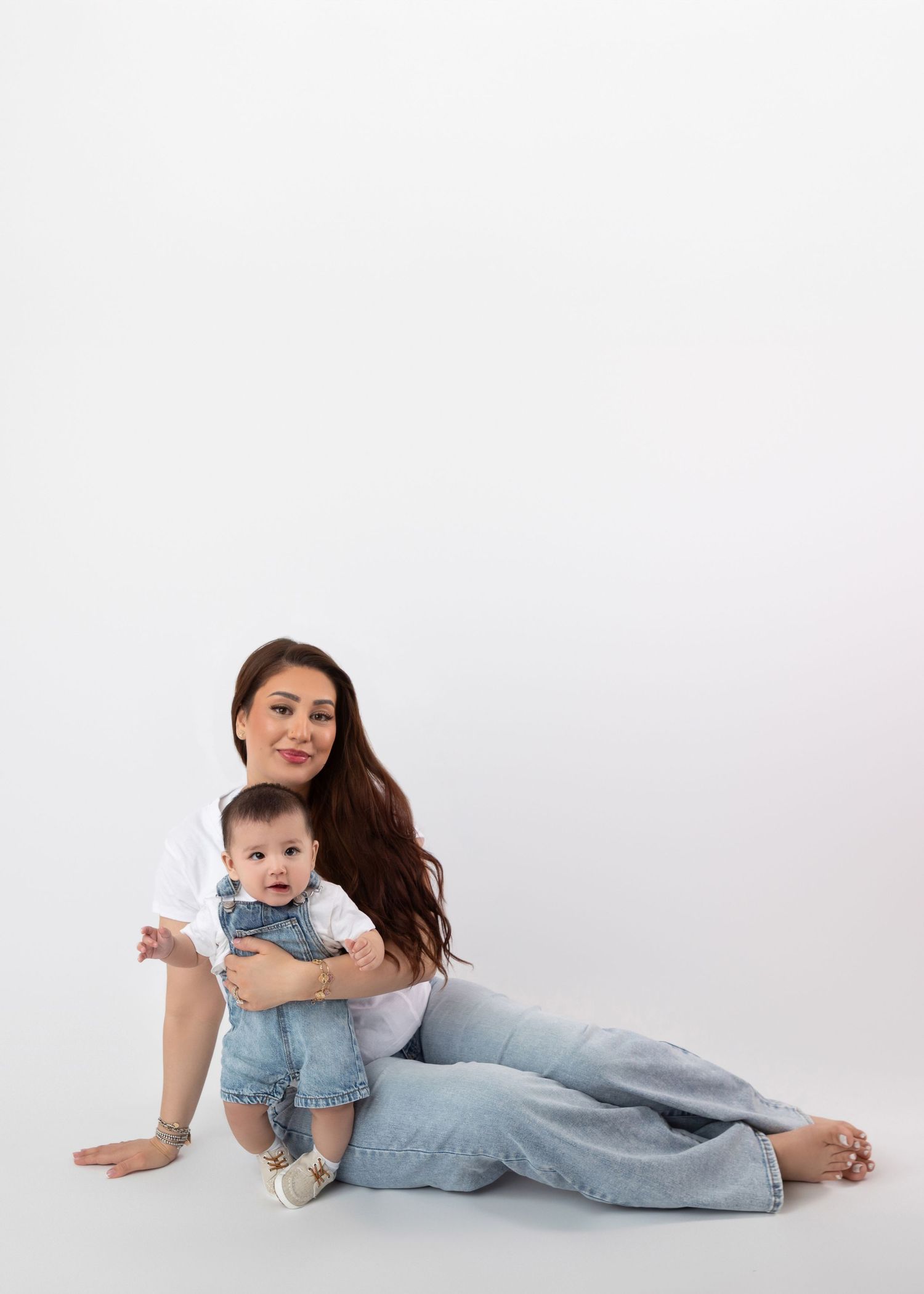 Mother in white top sitting on floor cradling baby in denim overalls.
