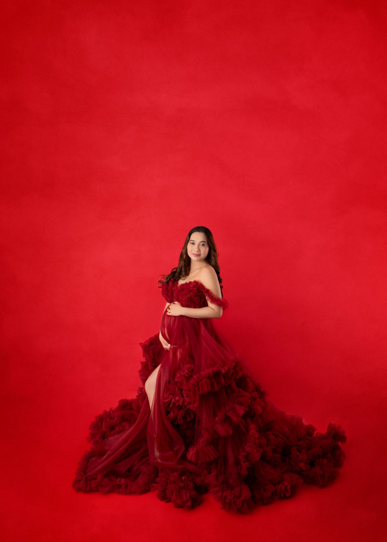 A pregnant woman in a stunning dark red ruffled tulle gown stands gracefully against a red backdrop.