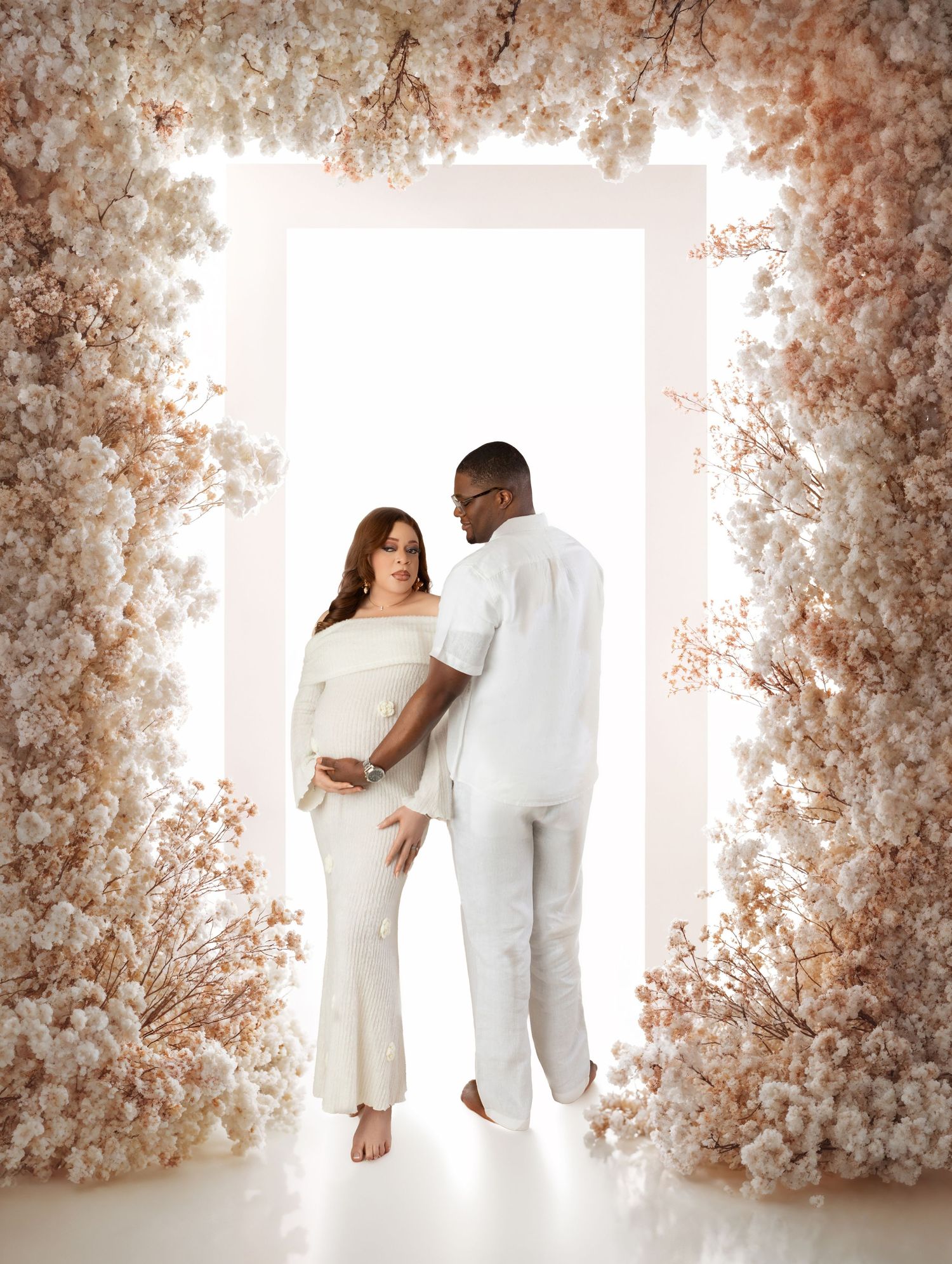 Expecting couple in white outfits standing in a glowing doorway framed by soft cream floral arrangements.