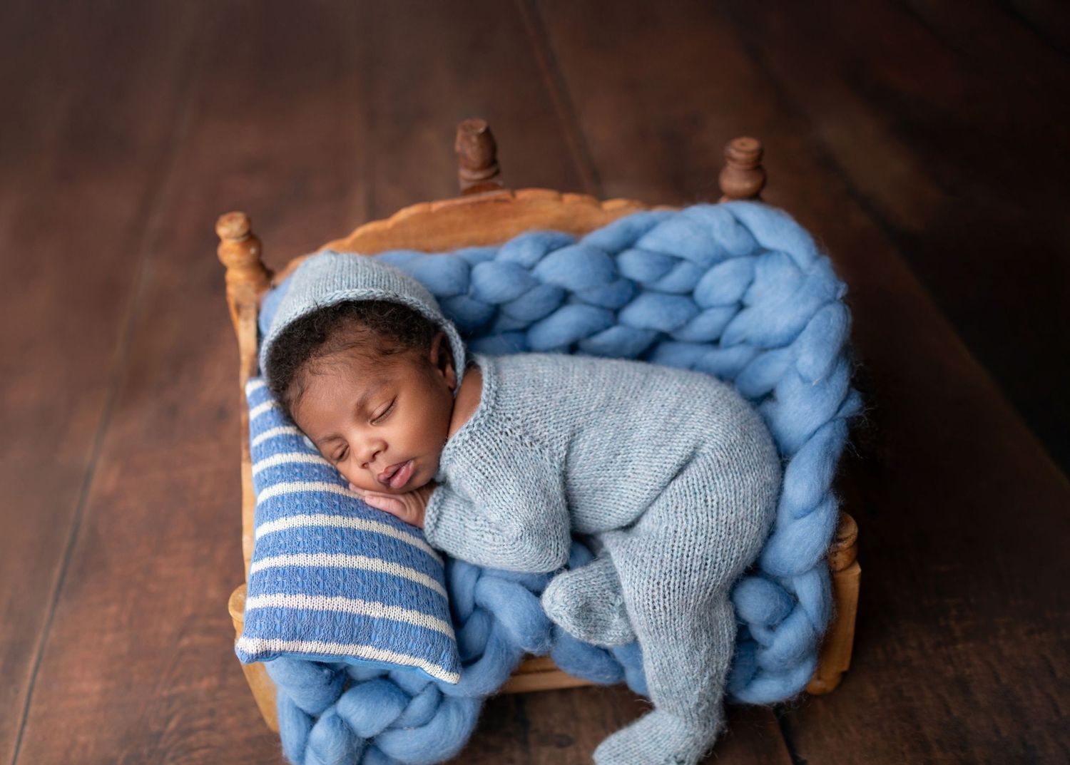 Newborn baby in blue knit outfit sleeping in wooden mini bed with chunky blue blanket.