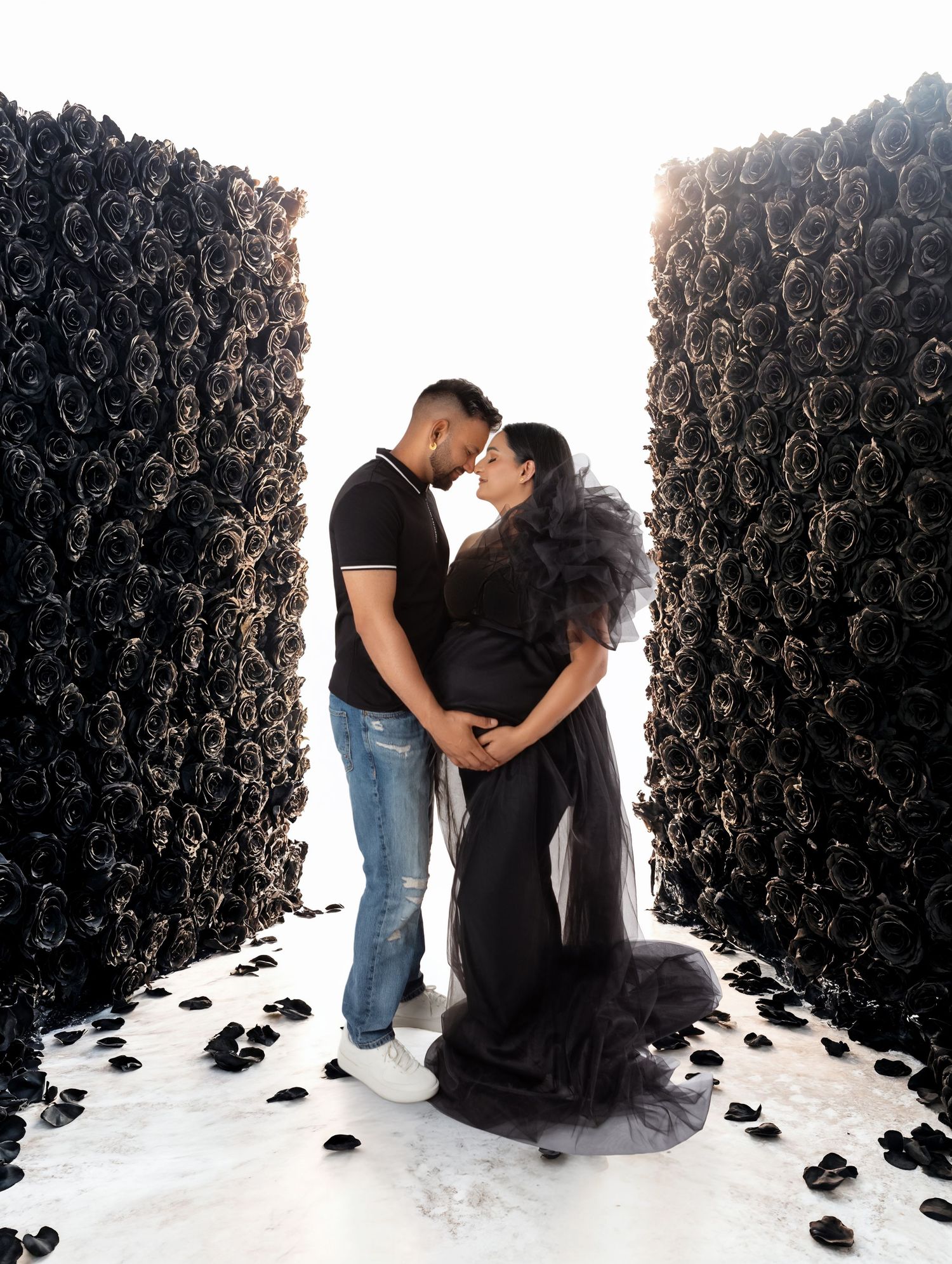 Couple in black outfits sharing a tender moment between walls of black roses in a dramatic maternity photo shoot.