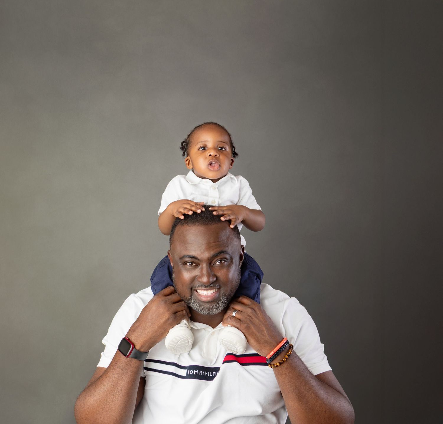 A smiling man in a white polo shirt carries a happy toddler on his shoulders against a gray background.