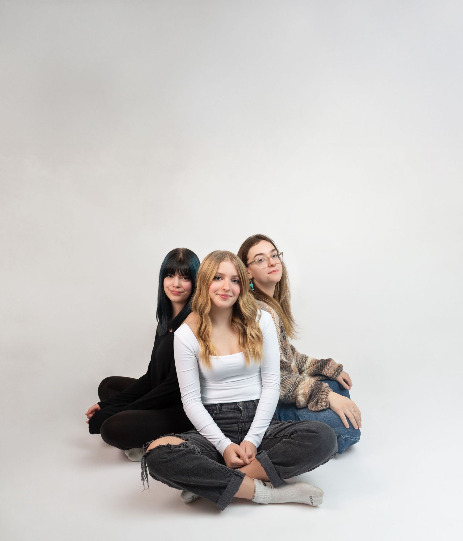 Three young women sitting cross-legged on white background, wearing casual outfits, smiling and posing together for a portrait.
