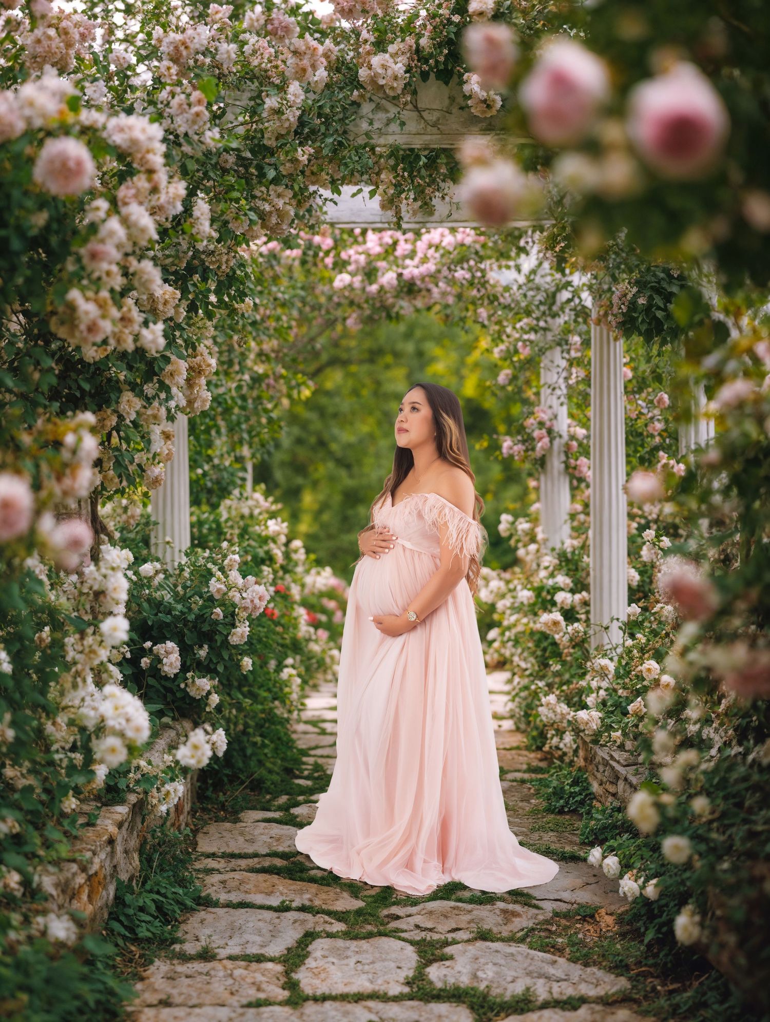 Pregnant woman in pink off-shoulder gown stands under floral archway in lush garden for maternity portrait.