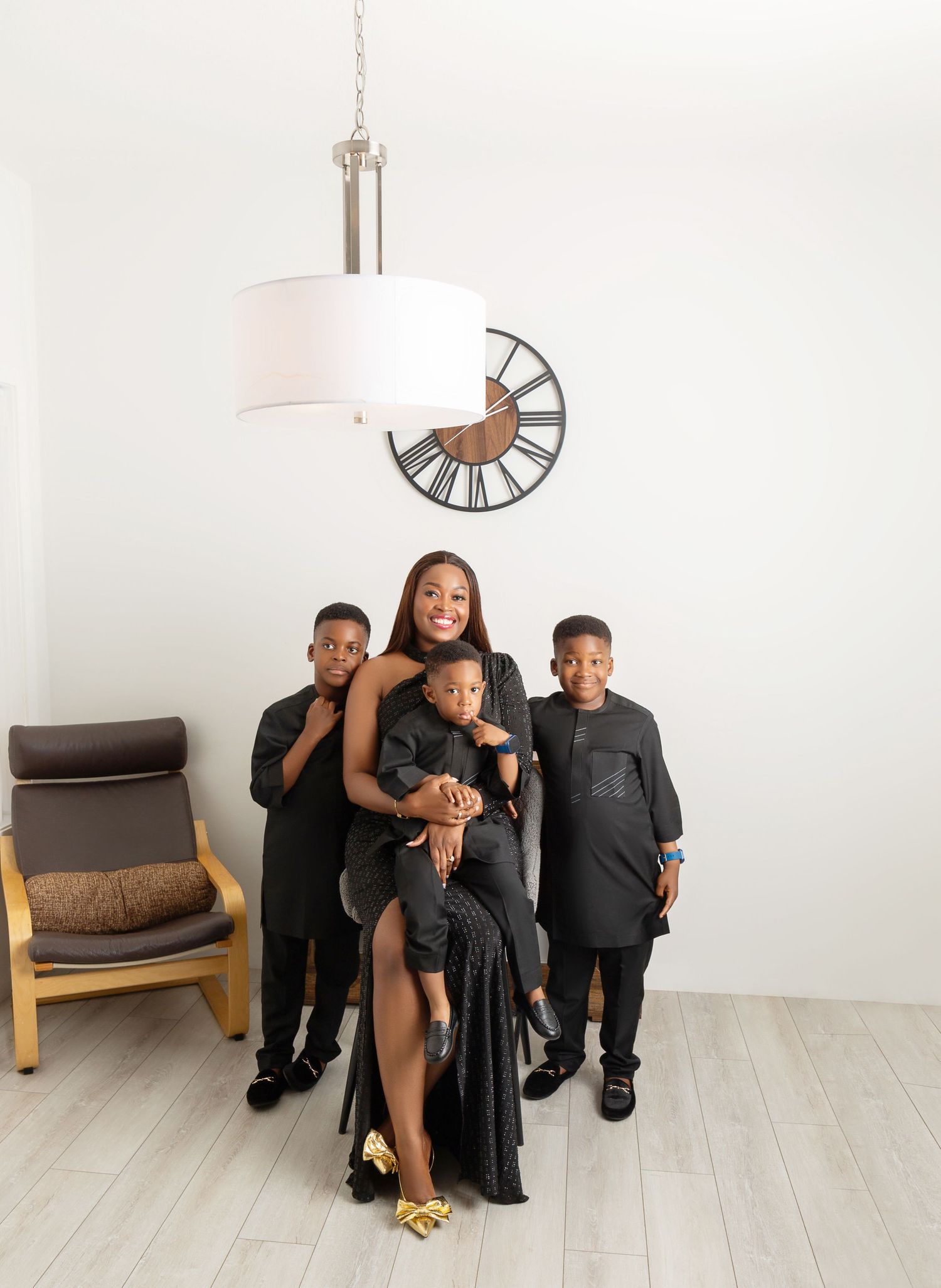 A woman in a black gown poses with three children in matching black outfits in a bright white room.