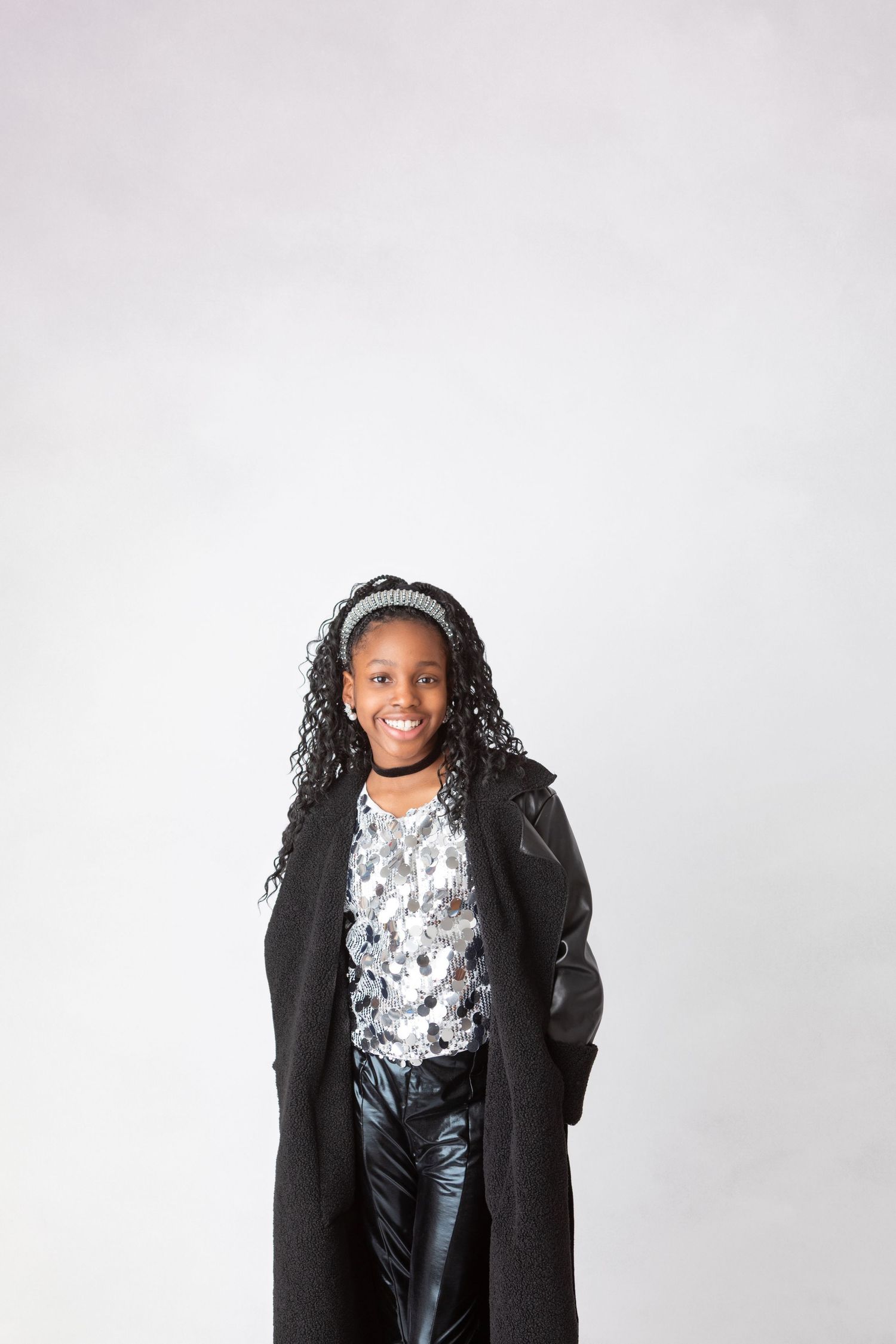 Stylish young Black girl in a long black coat, silver top, and leather pants, smiling confidently at camera.