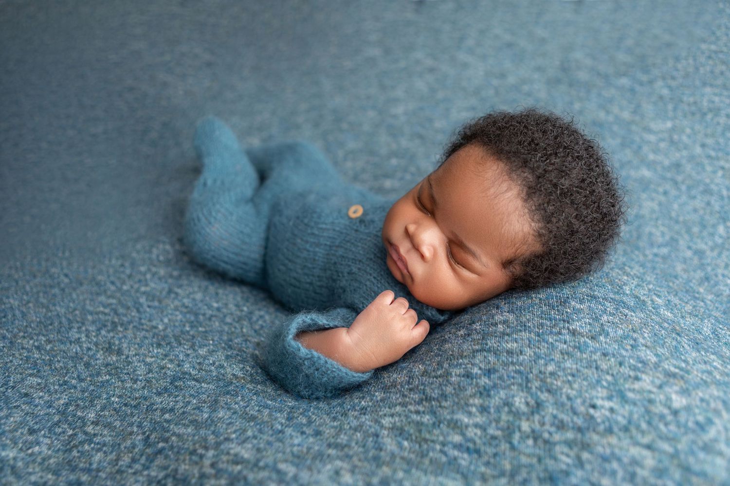 Sleeping newborn baby in teal knit romper posed on blue textured backdrop in professional newborn photography.