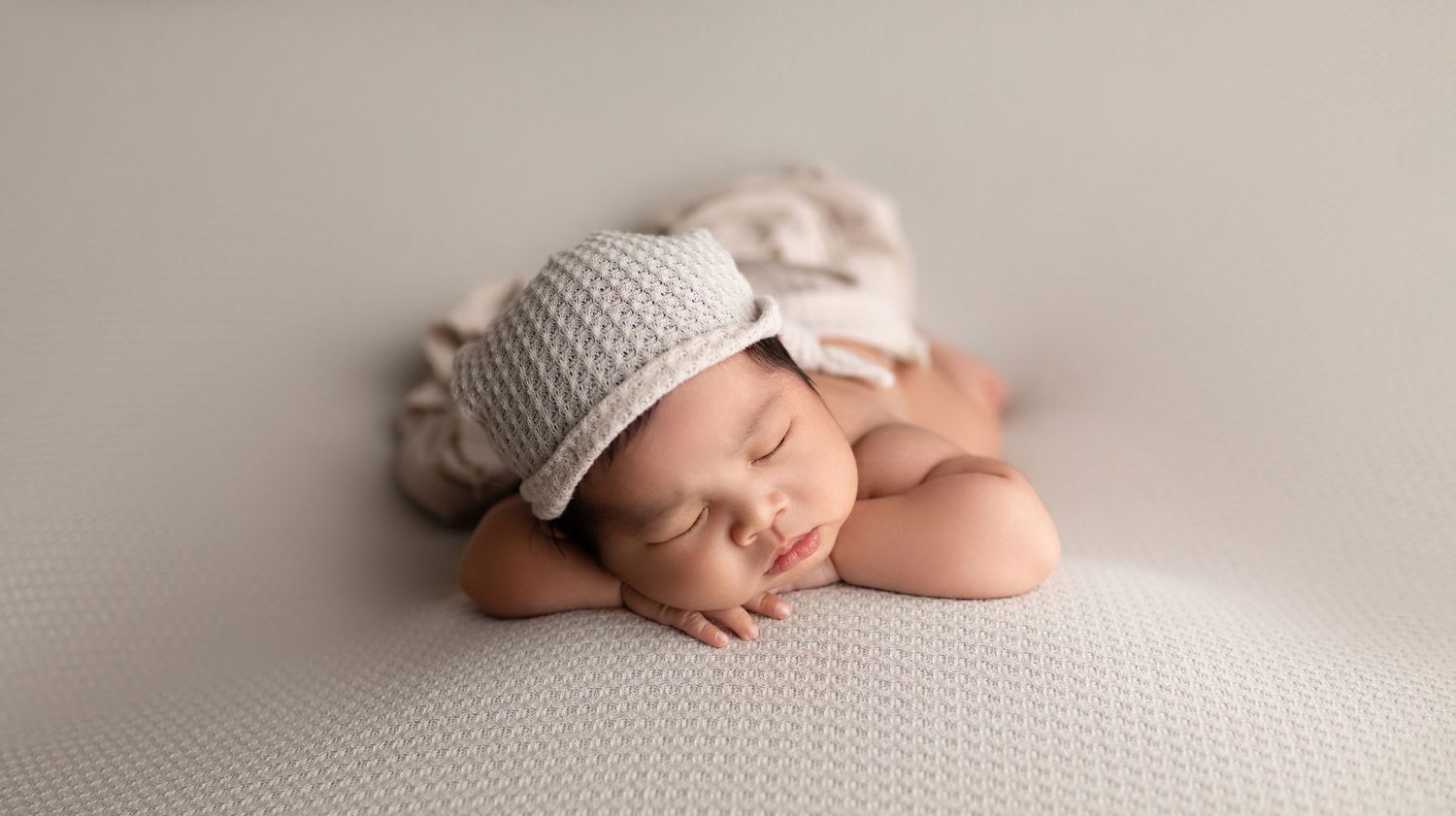 Peaceful newborn baby sleeping curled up wearing a soft gray knitted hat on a light gray textured background.