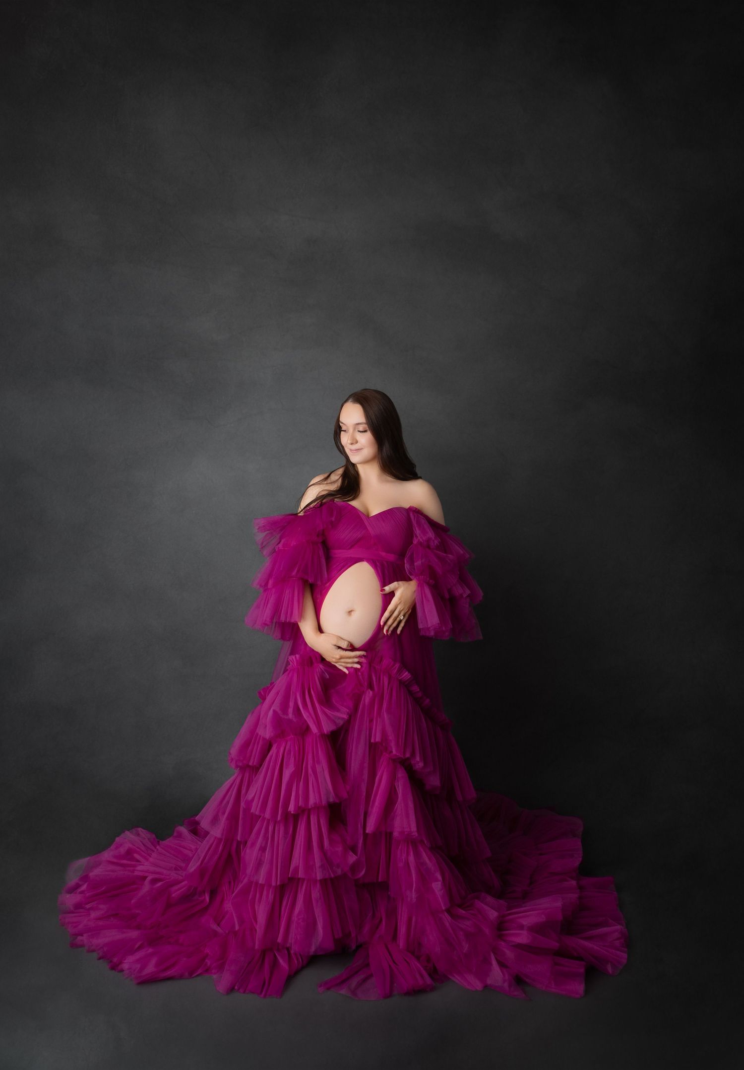 Expecting mother in bold magenta off-shoulder ruffled tulle gown poses against dark gray studio backdrop.