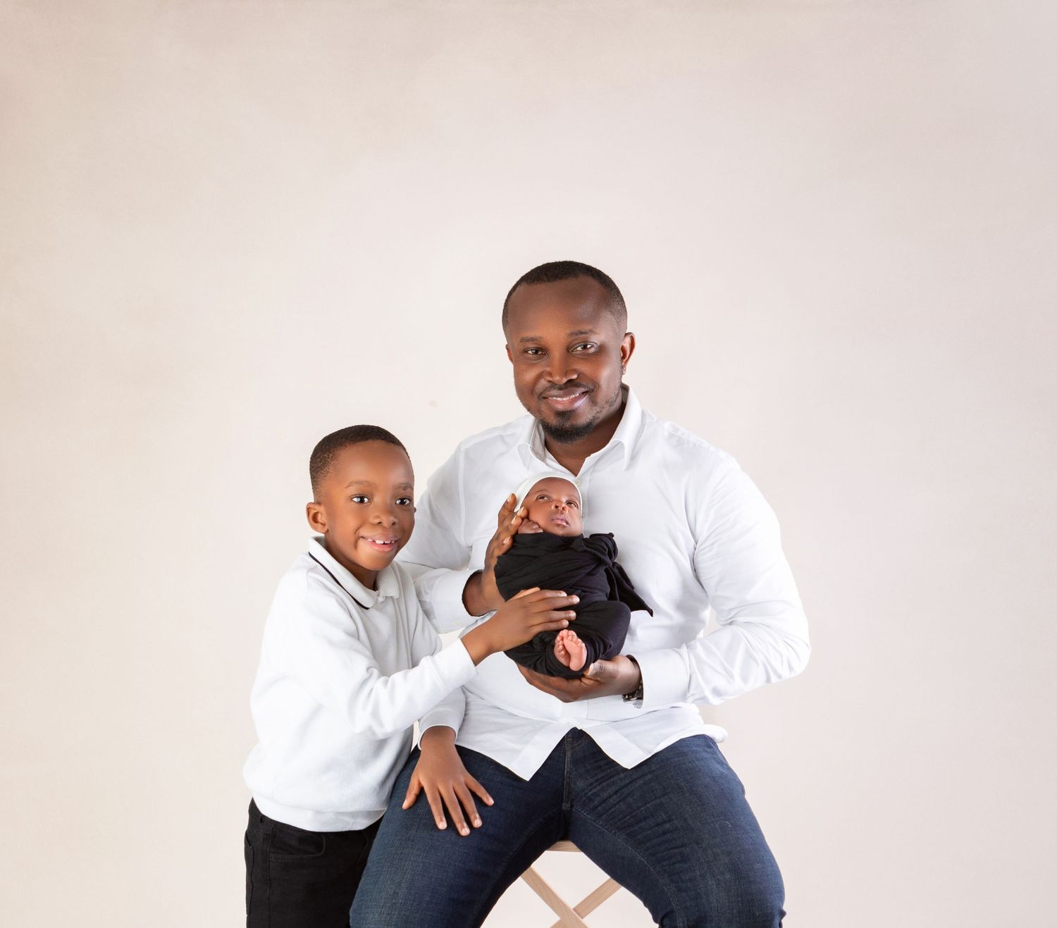 A father in a white shirt holds his swaddled newborn baby while his young son smiles beside him in a studio photo.