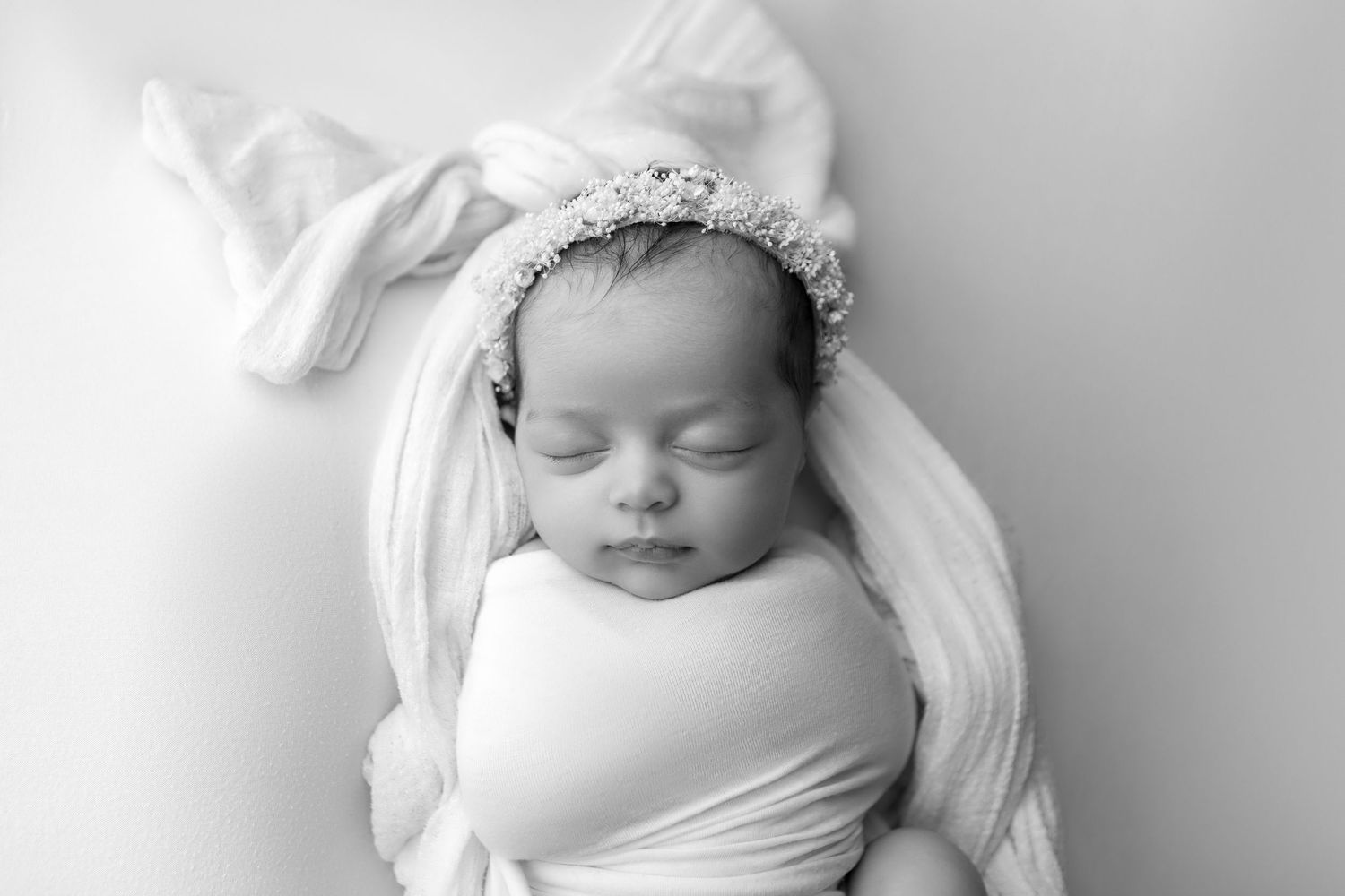 Black and white portrait of swaddled newborn with bow headband sleeping peacefully.
