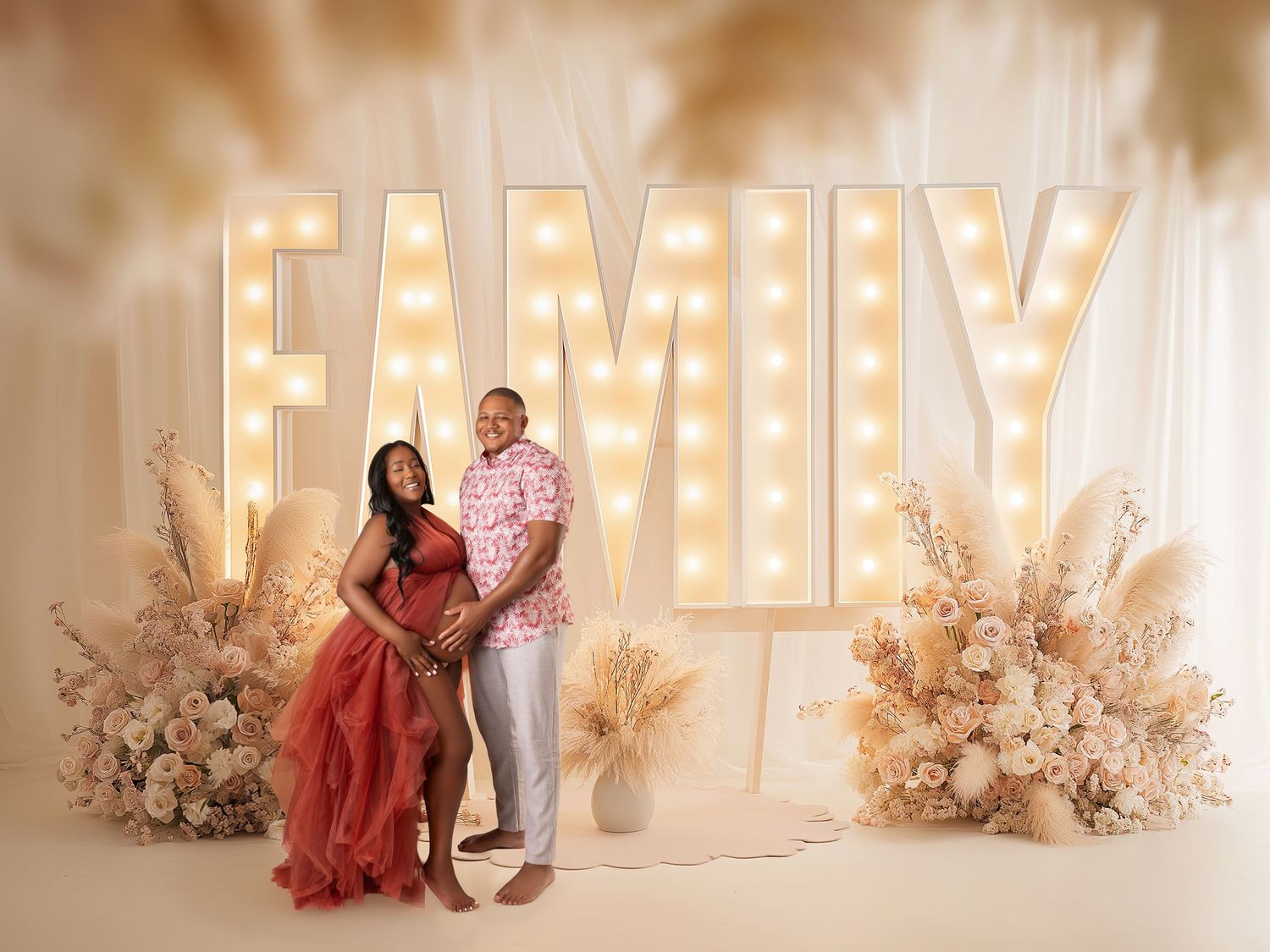 Couple poses before illuminated FAMILY marquee letters with floral arrangements in a warm maternity photo shoot.