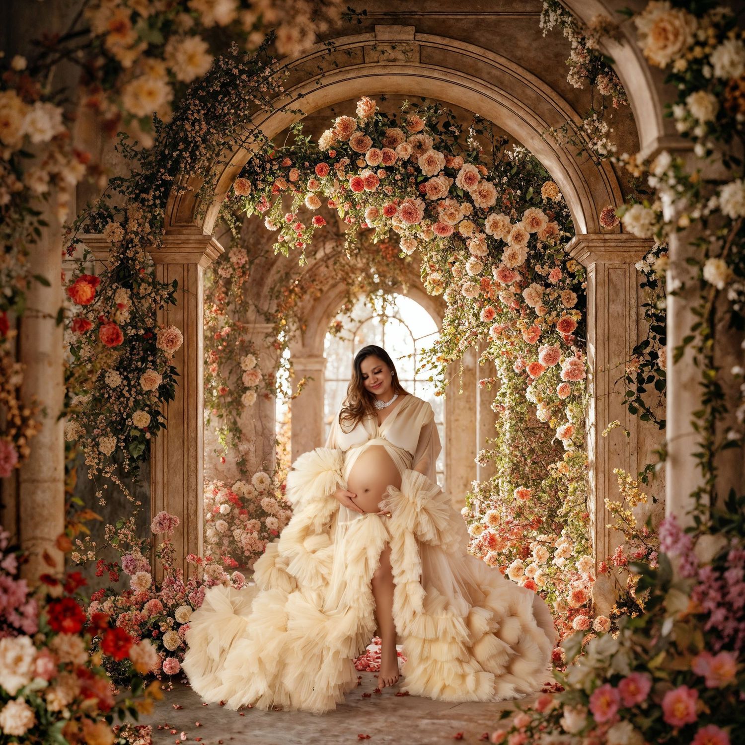 Pregnant woman in cream ruffle gown standing under floral arch with roses in dreamy studio setting.